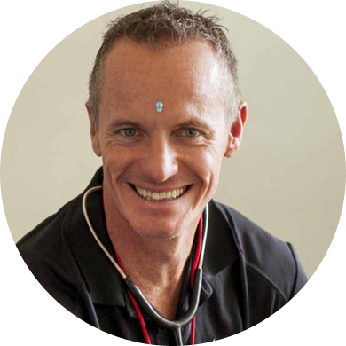 A smiling man with short gray hair and a small decorative mark on his forehead, wearing a black shirt and a stethoscope around his neck.