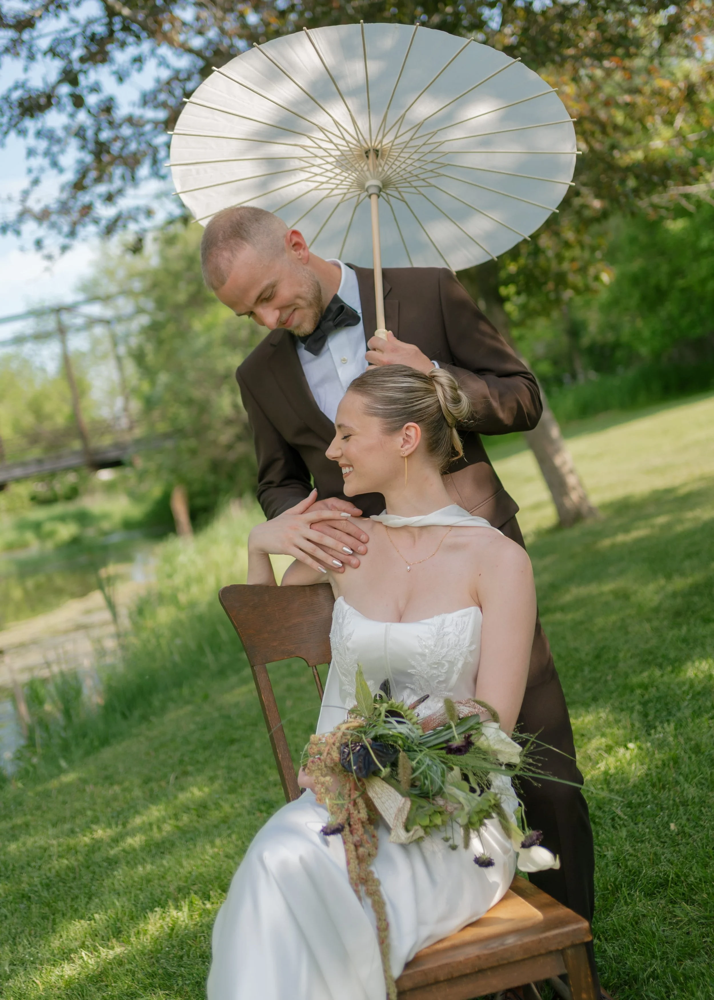 A man in a tuxedo holding a white parasol over a woman dressed in a white wedding gown, sitting on a wooden chair outdoors on a sunny day.