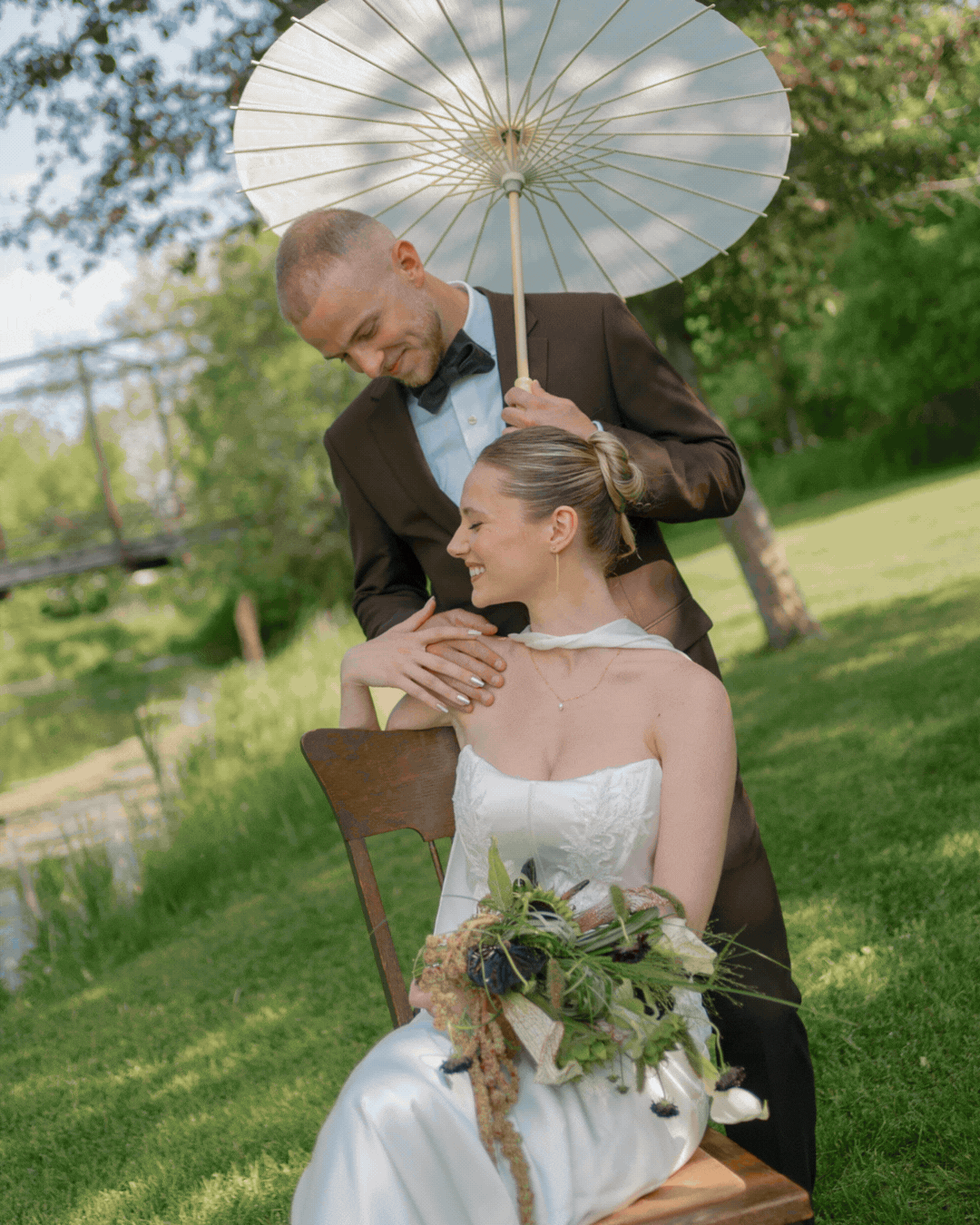 A groom in a brown suit and black bow tie is holding a white parasol over a seated bride in a white wedding dress with a bouquet, both smiling outdoors on a grassy day with trees in the background.