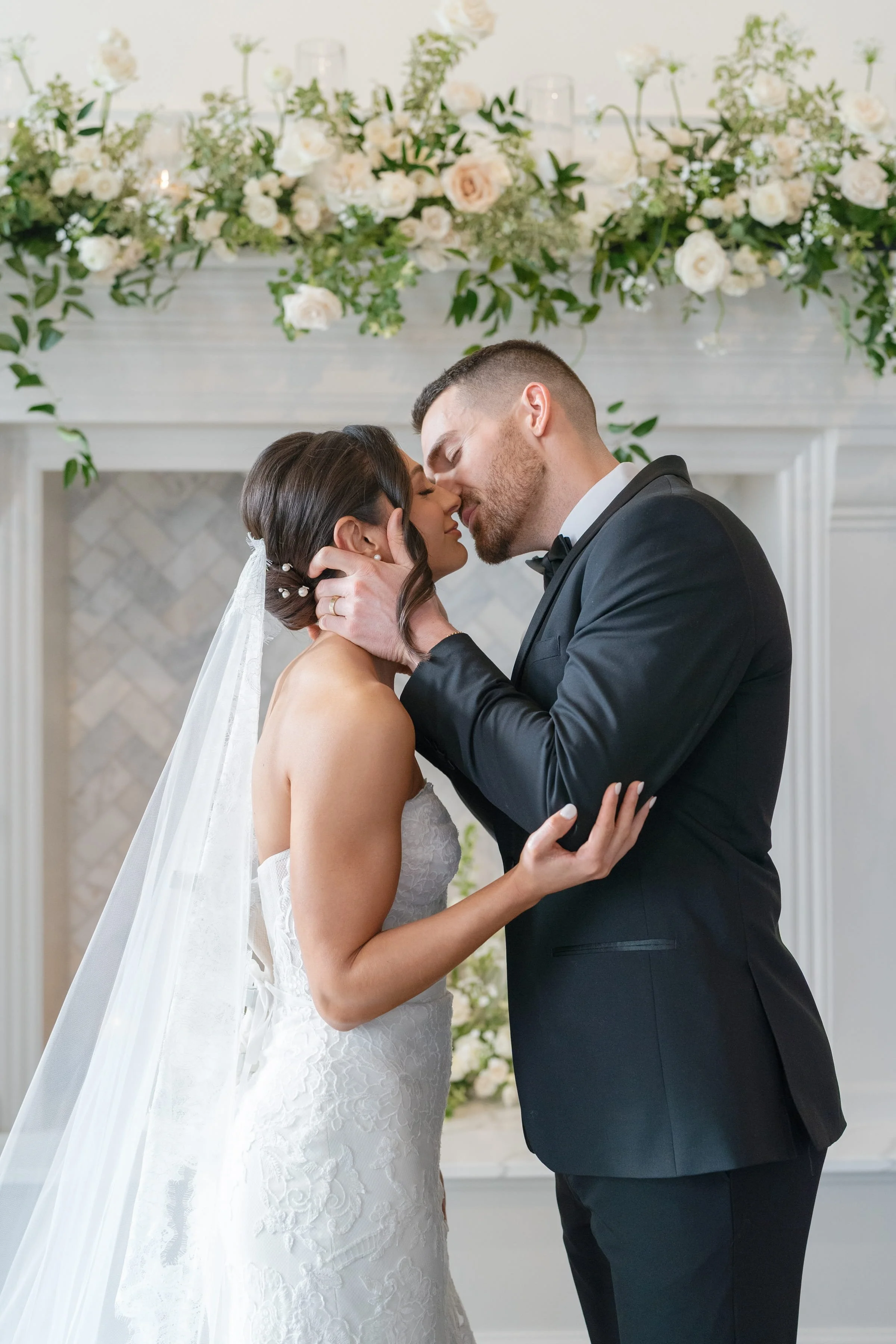 A bride and groom share a kiss during their wedding ceremony in an elegant decorated room with floral arrangements and candles.