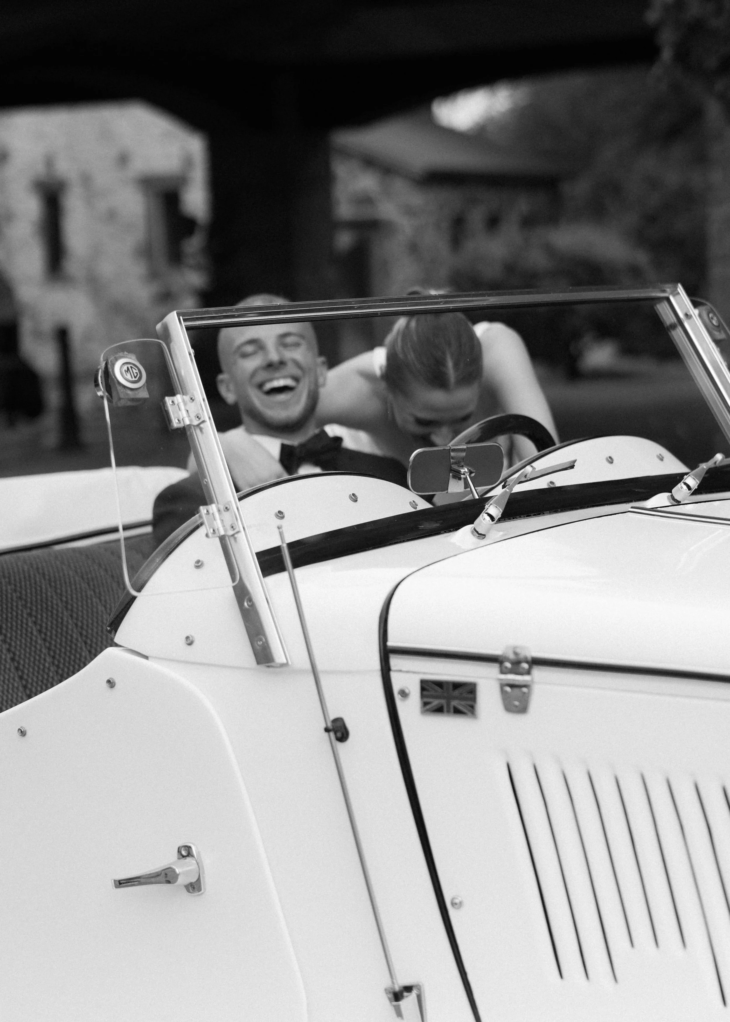 A black and white photo of a smiling man and a woman in a vintage convertible car. The car has a small Union Jack flag on the side, and the man appears to be driving. The background shows trees and a building, possibly in an outdoor setting.