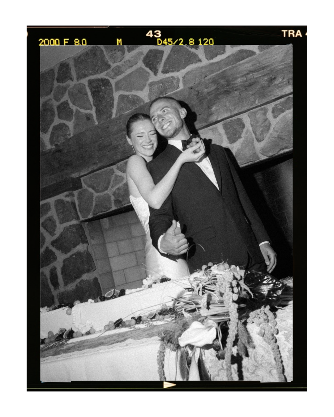 Black and white photo of a happy bride and groom smiling and hugging at their wedding reception, with a decorated wedding cake in the foreground and a brick and stone fireplace in the background.