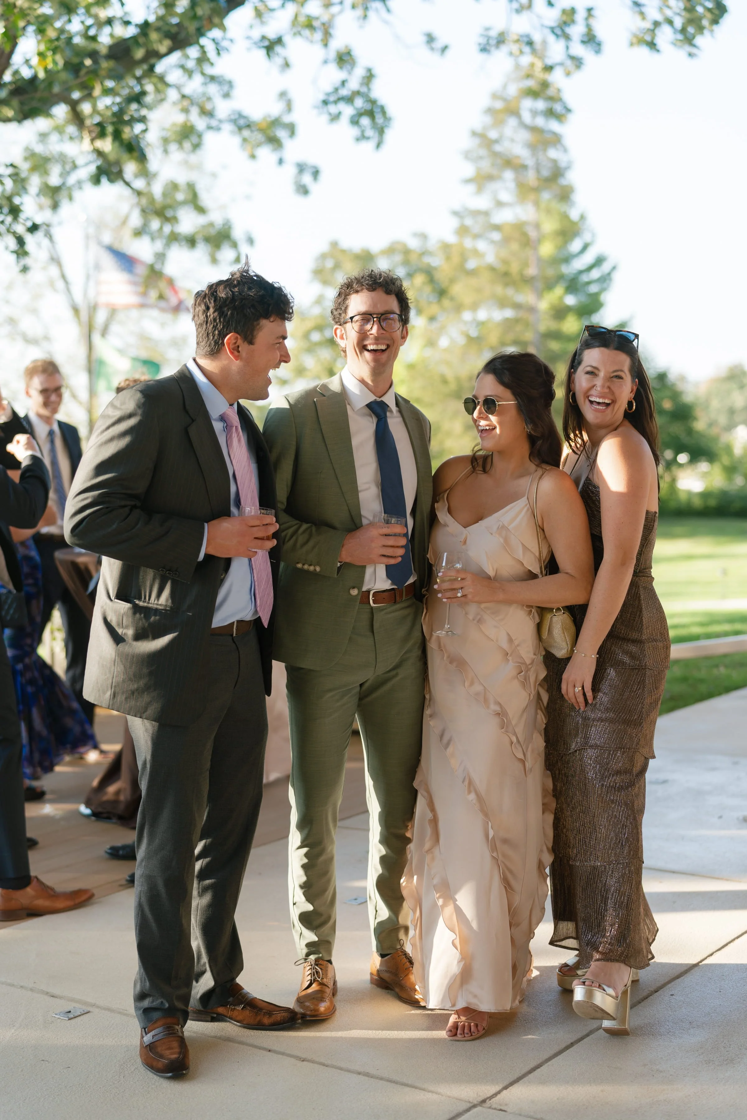 A group of five people, three men and two women, wearing formal attire, are standing outdoors on a sunny day, laughing and enjoying each other's company. The background shows other people and trees with an American flag.