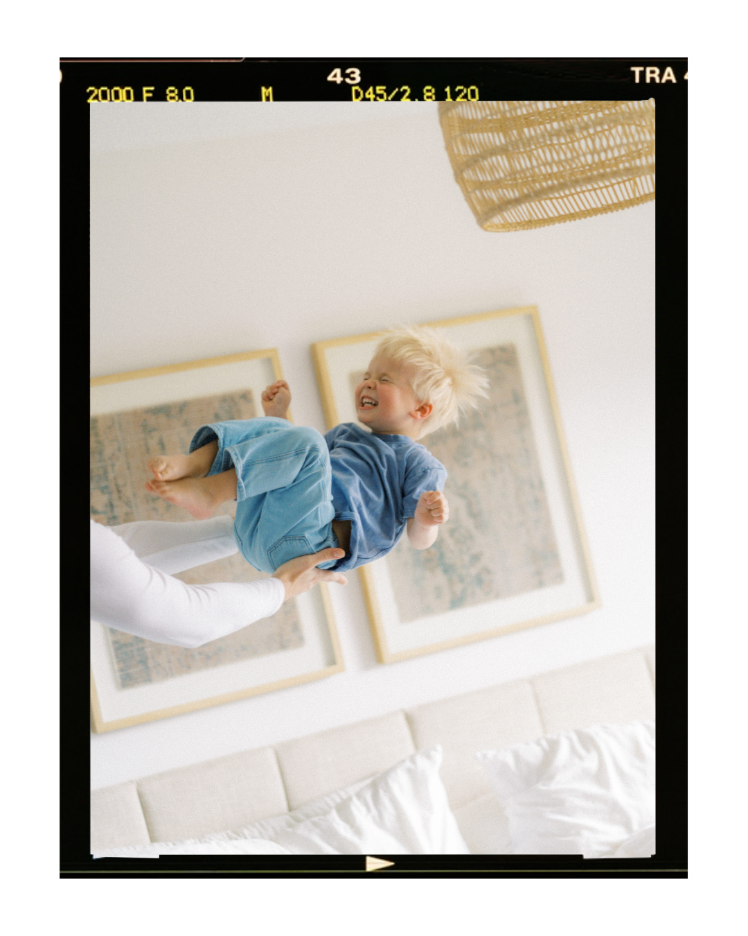 A young boy with blonde hair laughing and smiling while being thrown in the air indoors, with framed art on the wall behind him.