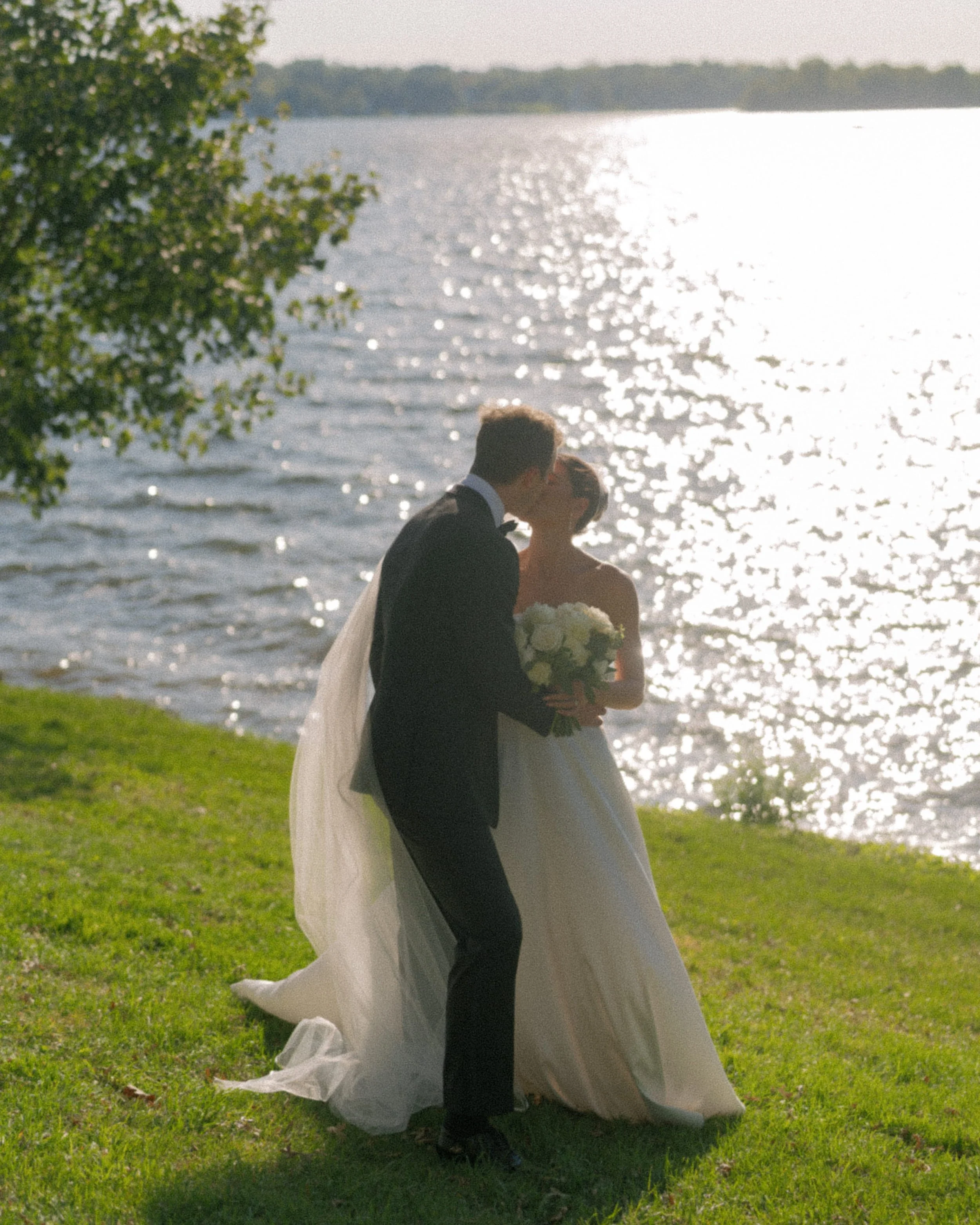 A bride and groom share a kiss by a lakeside on a sunny day, with sunlight reflecting on the water and lush green grass in the foreground.