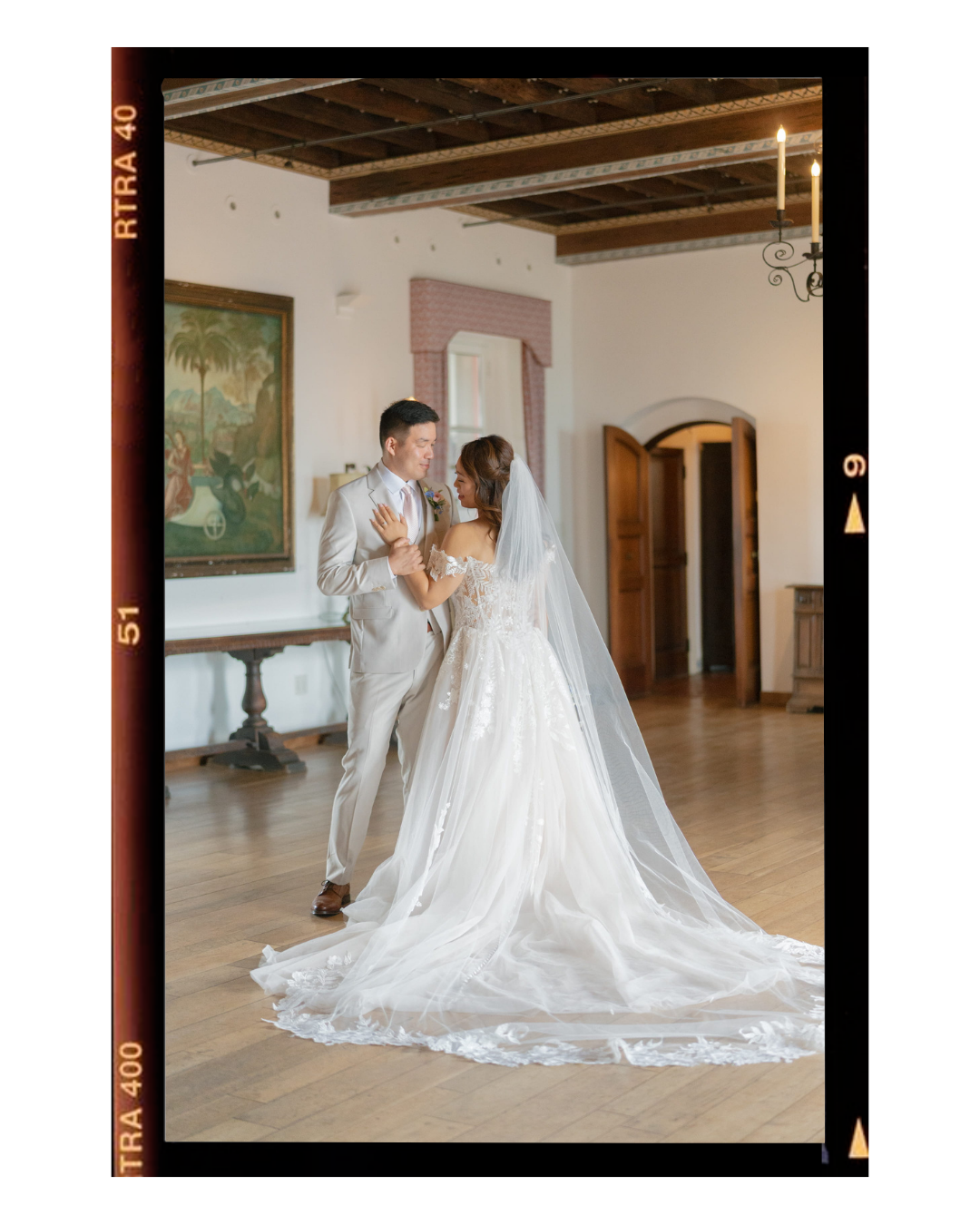 A bride and groom dancing indoors, with the bride wearing a white wedding gown and veil, and the groom in a beige suit. The room has wooden floors, a painting on the wall, and wooden doors.