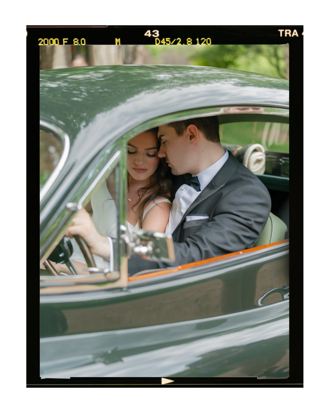 A newlywed couple in wedding attire sitting inside a vintage car, close together and sharing an intimate moment.