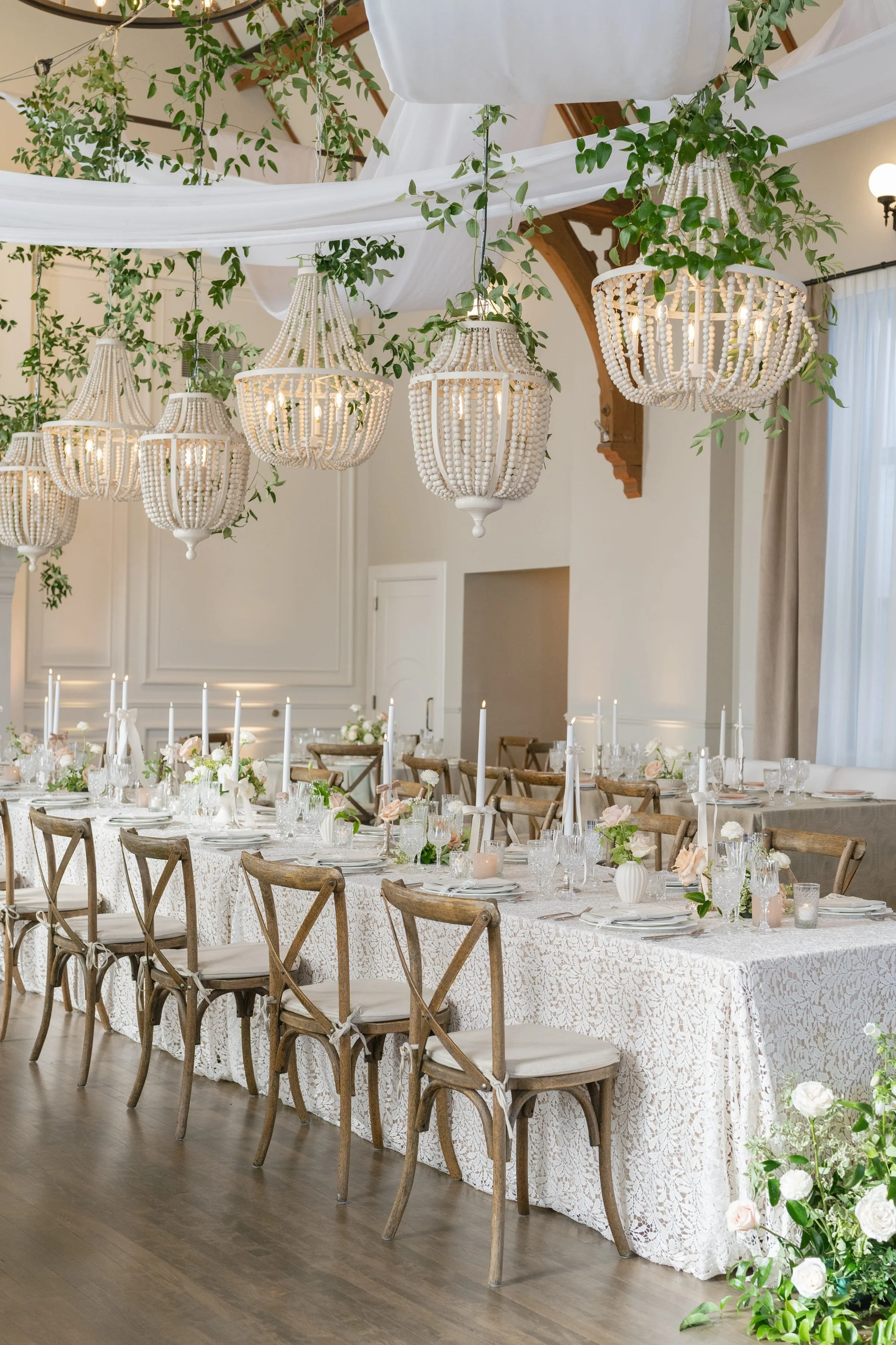 Elegant indoor wedding reception with a long decorated table, wooden chairs, white lace tablecloth, floral centerpieces, and chandeliers adorned with greenery hanging from the ceiling.
