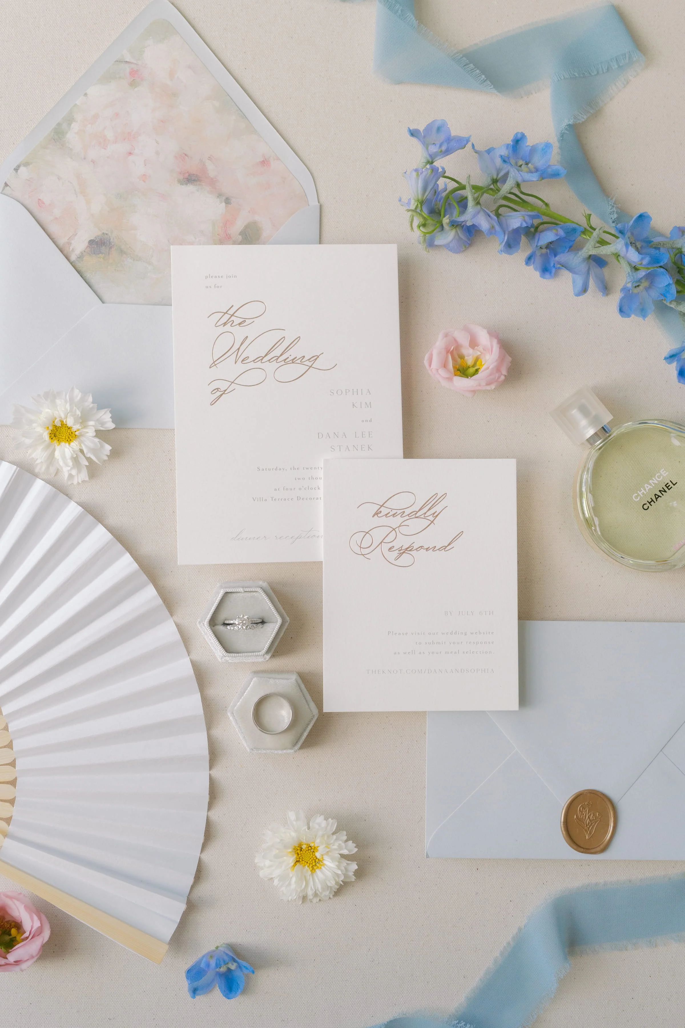 Wedding invitation suite with floral accents, a white fan, a blue ribbon, and a Chanel perfume bottle.