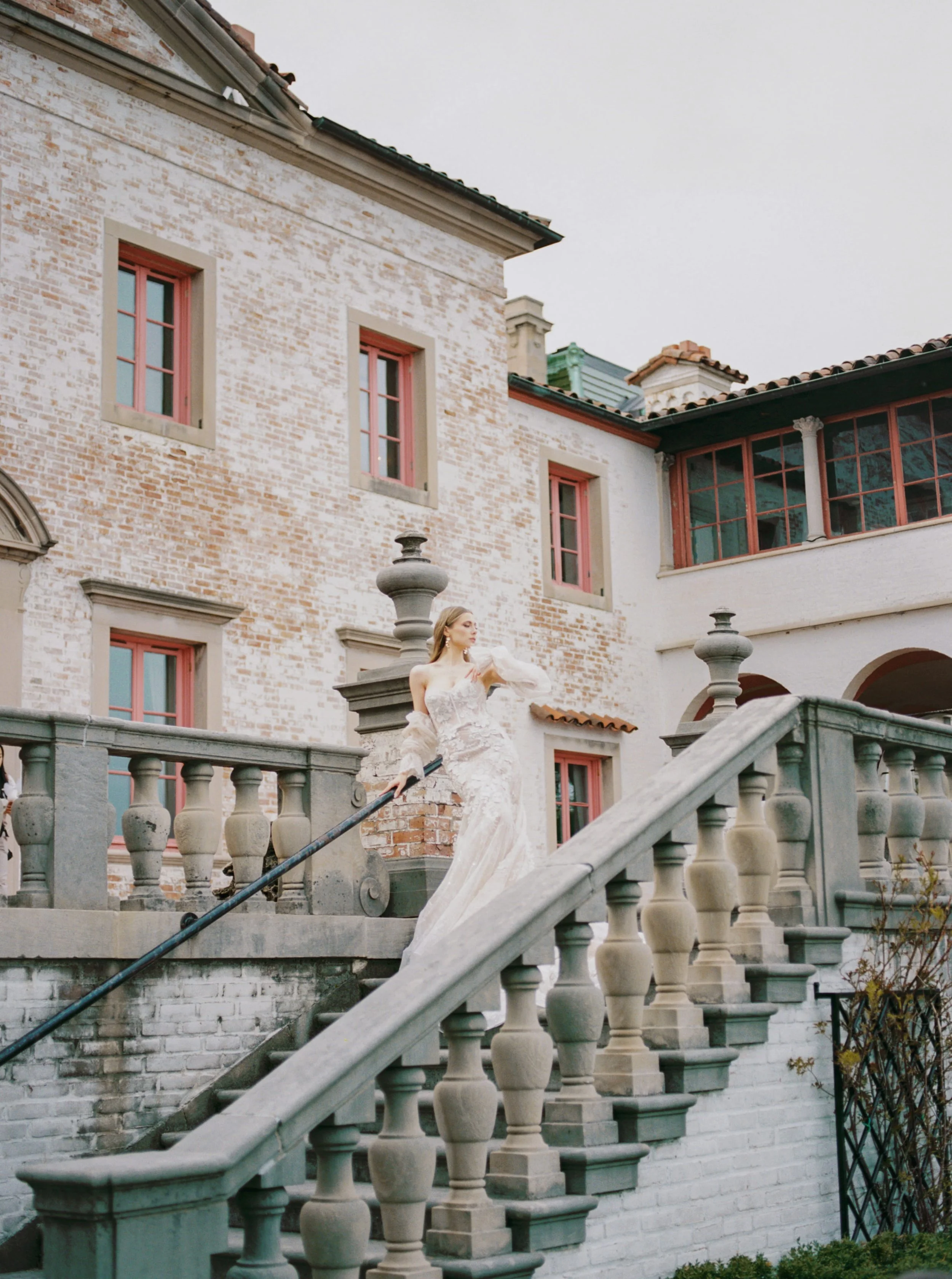 A woman in a white dress stands on an outdoor staircase with a stone railing, in front of a historic brick building with pink window frames and a tiled roof.