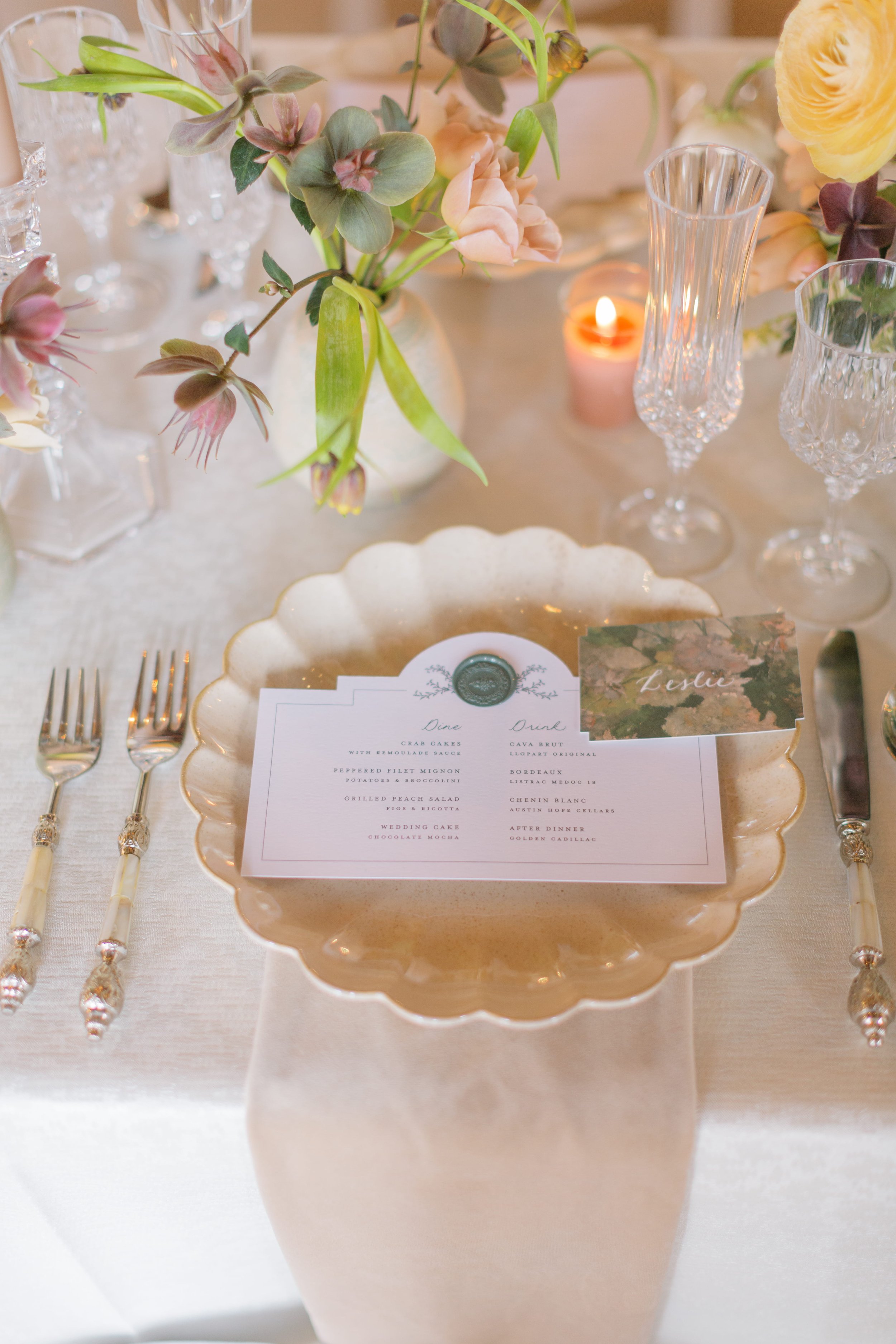 Elegant table setting with a white vase of pink and green flowers, crystal glasses, a lit candle, a paper menu inside a scalloped gold-edged white dish, and a place card with the name 'Leslie' on a wedding or formal event table.
