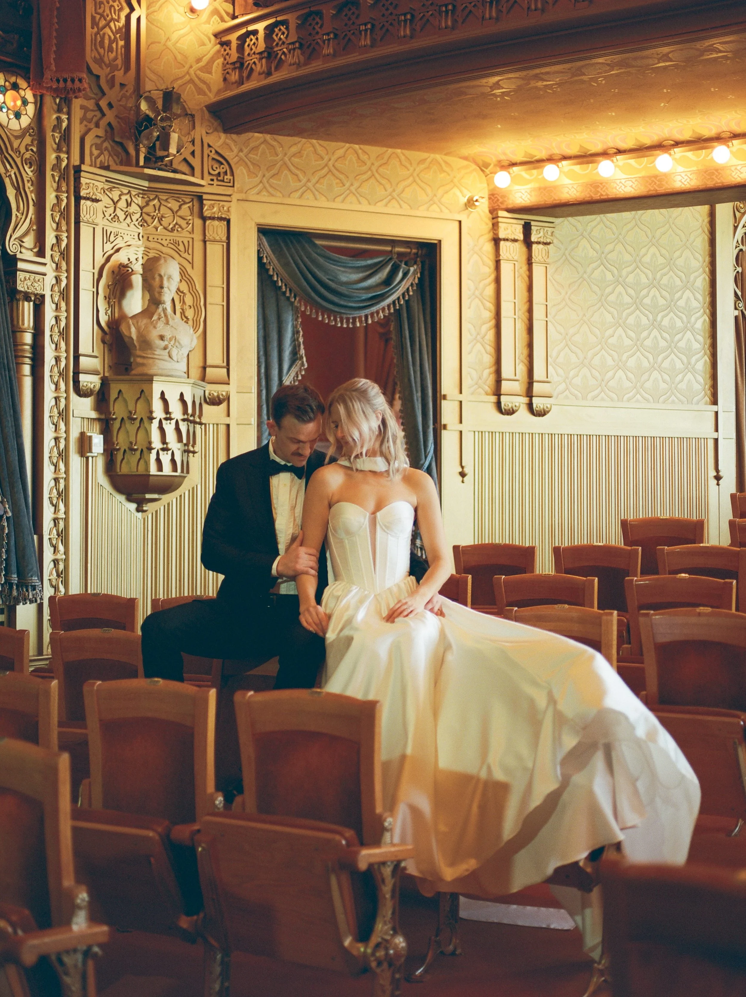 A man and woman dressed in formal attire, sitting closely together in an elegant, ornately decorated room with wooden chairs and gold accents, likely for a wedding or special event.