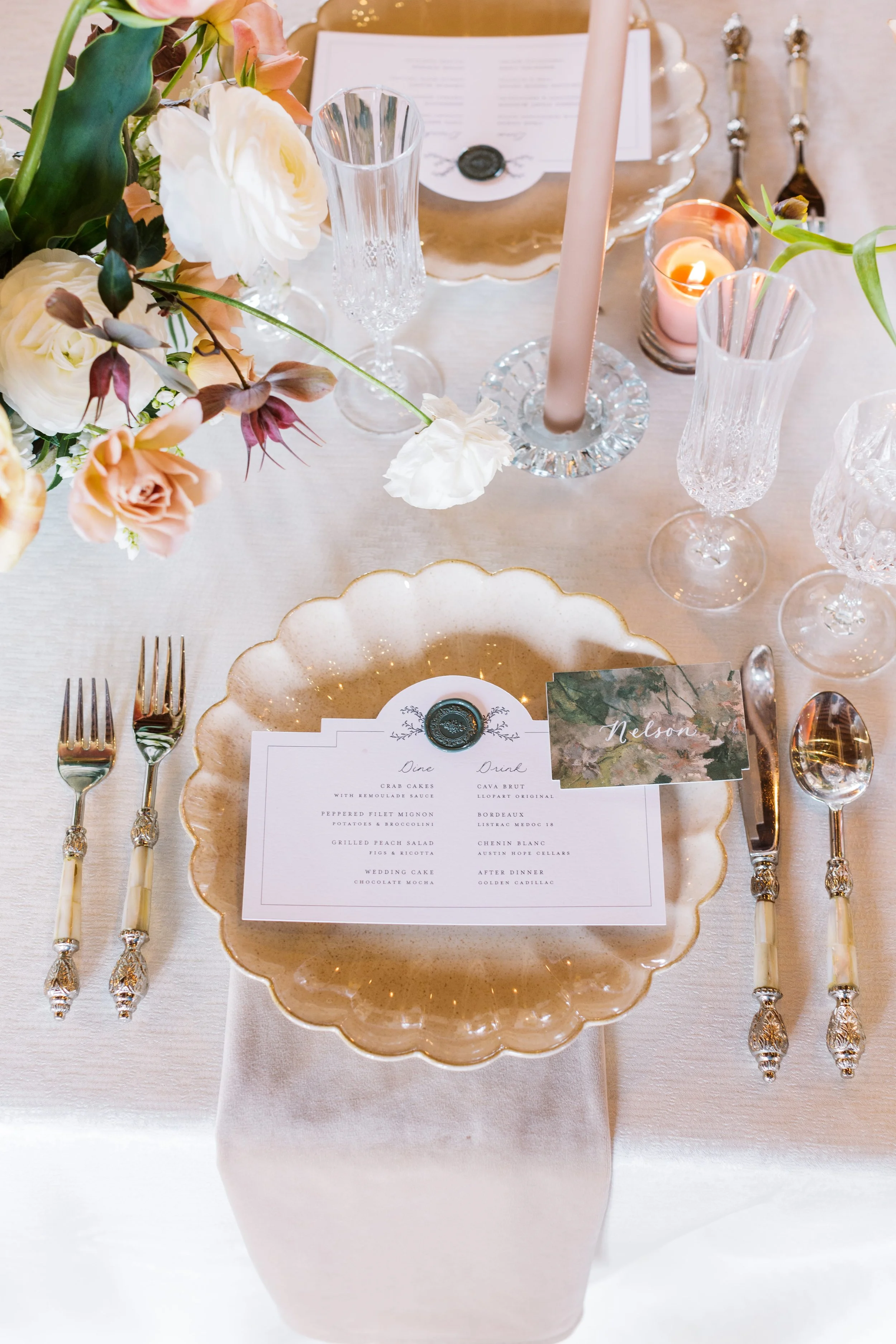 Elegant table setting with a floral centerpiece, crystal glasses, candles, and a printed menu on a gold-rimmed plate, all arranged around a white tablecloth.