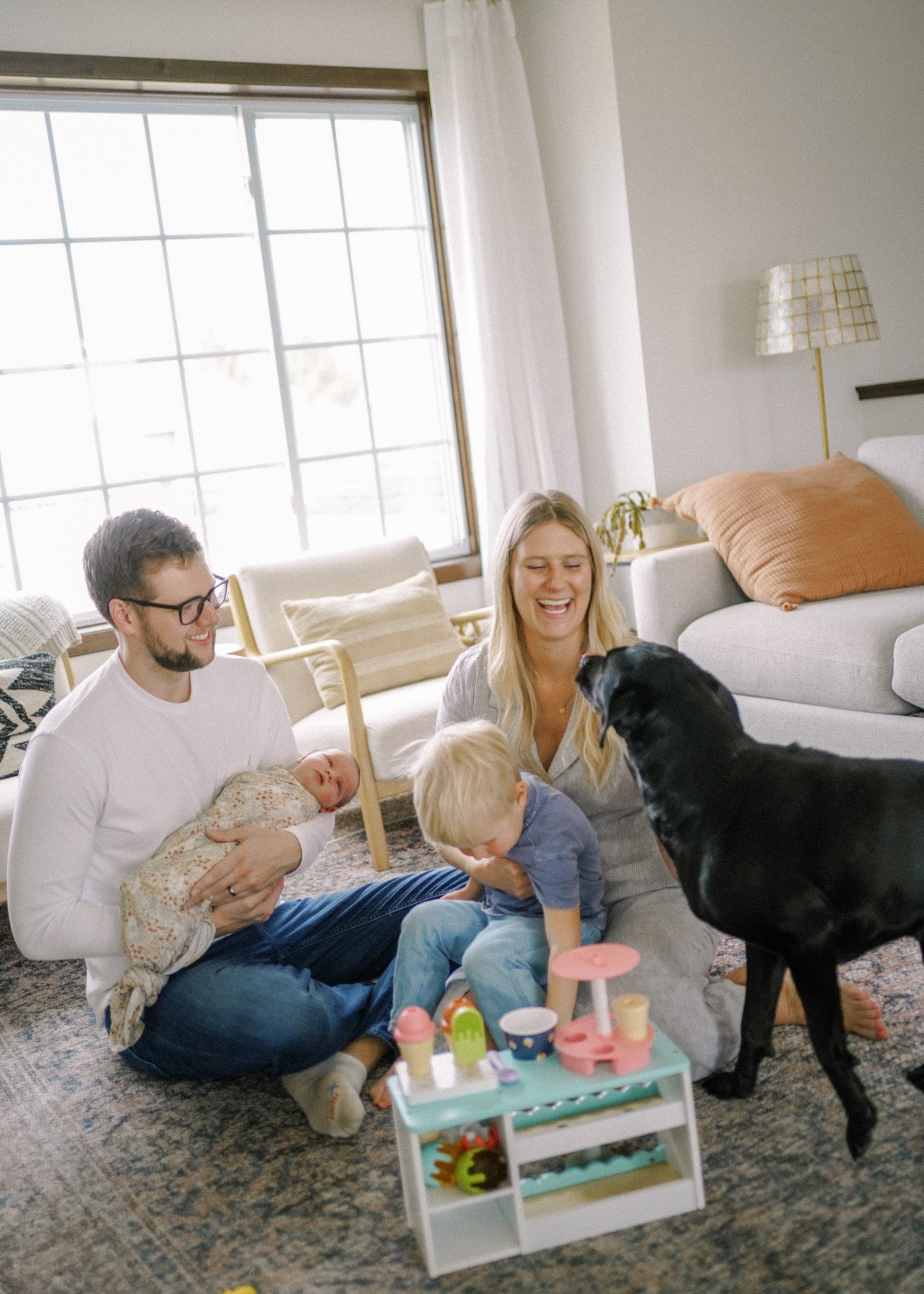 A family of five, including two adults, a baby, and two young children, enjoying a playful moment with their dogs in a bright living room. A white toy cart with colorful toy ice cream cones is in the foreground.
