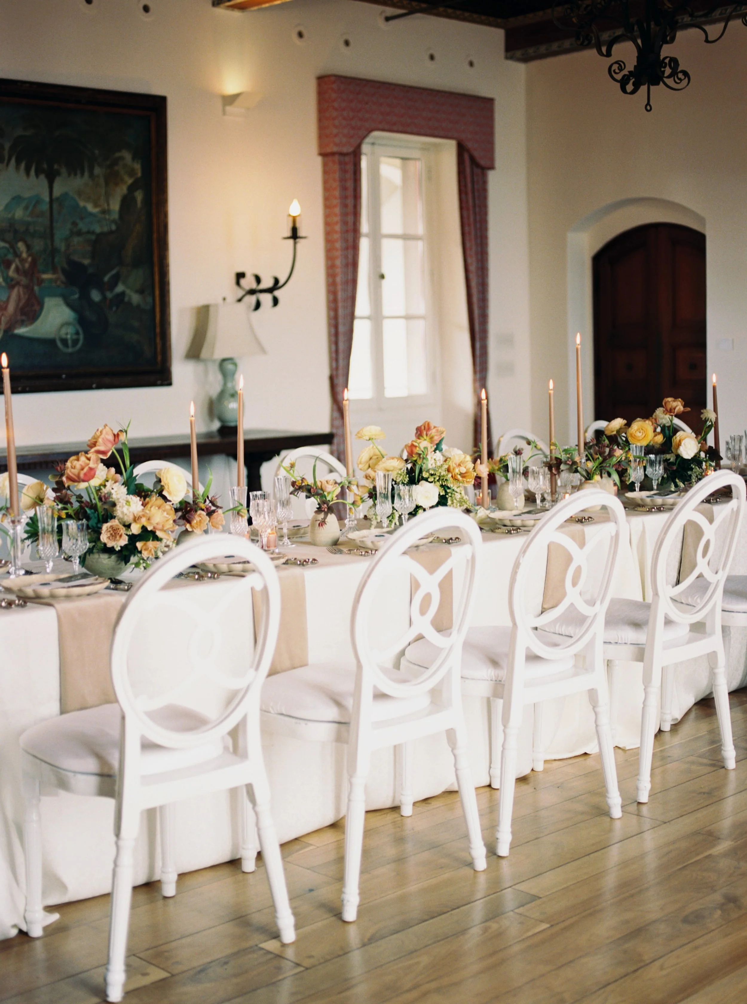 Elegant dining table set for a formal event with white chairs, floral arrangements, tall candles, and a long tablecloth in a room with wooden floors, painted wall, decorative curtains, painting, and vintage style lighting fixtures.
