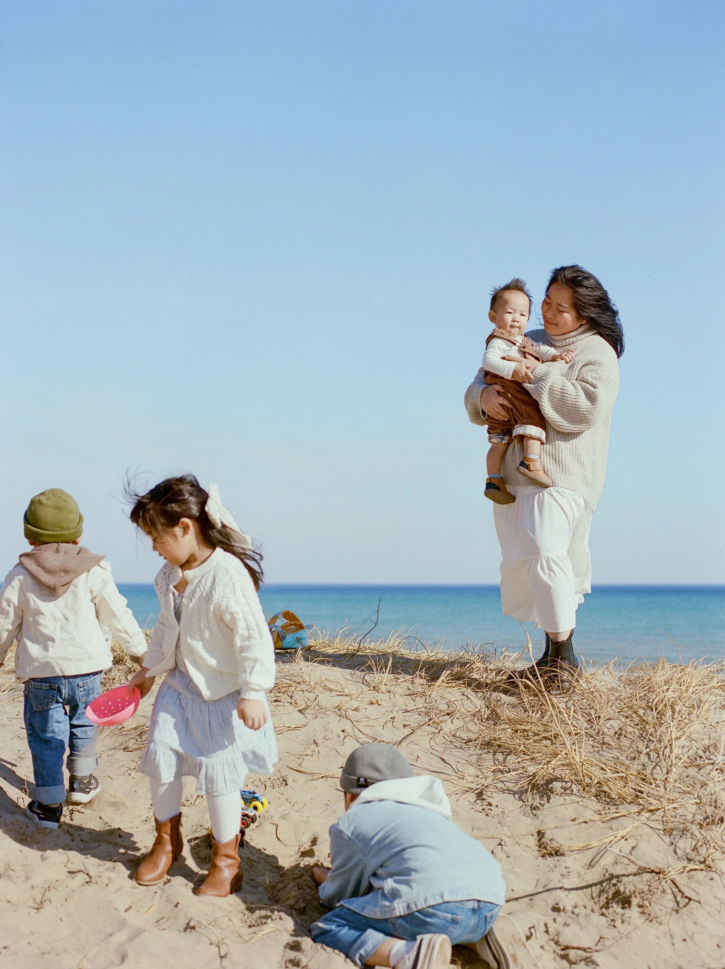 A group of children and a woman on a sandy beach with the ocean in the background, engaging in activities such as digging in the sand and holding a child.