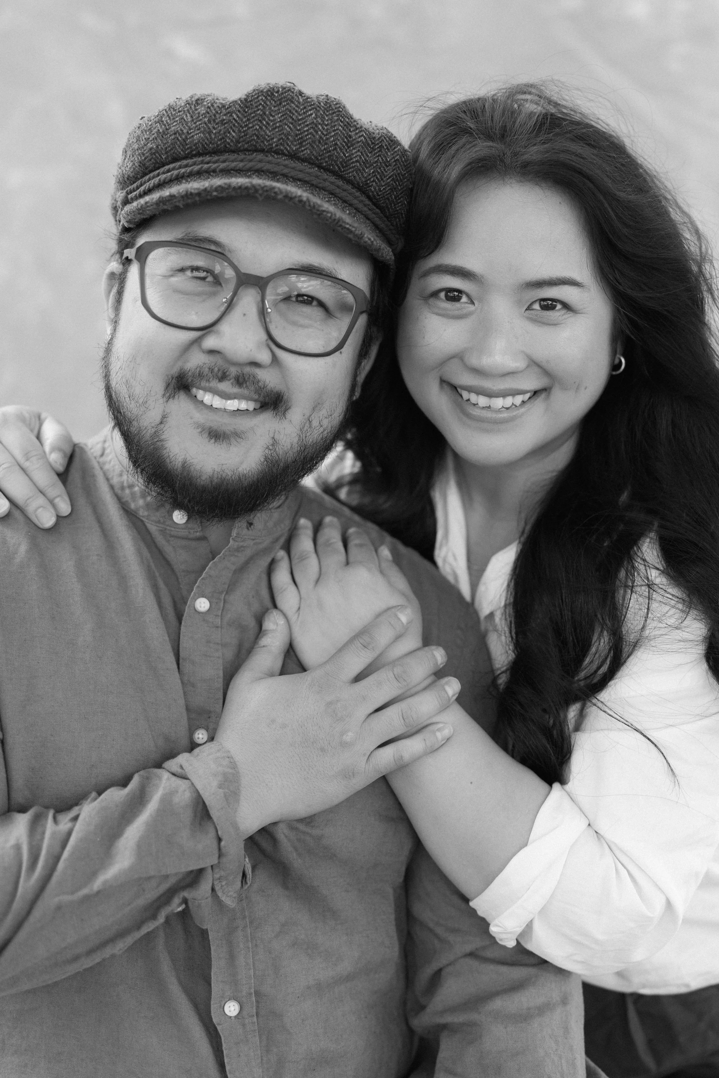 A black-and-white photo of a smiling man and woman embracing each other. The man is wearing glasses, a hat, and a button-up shirt. The woman has long, wavy hair and is wearing a light-colored blouse. They appear happy and close.