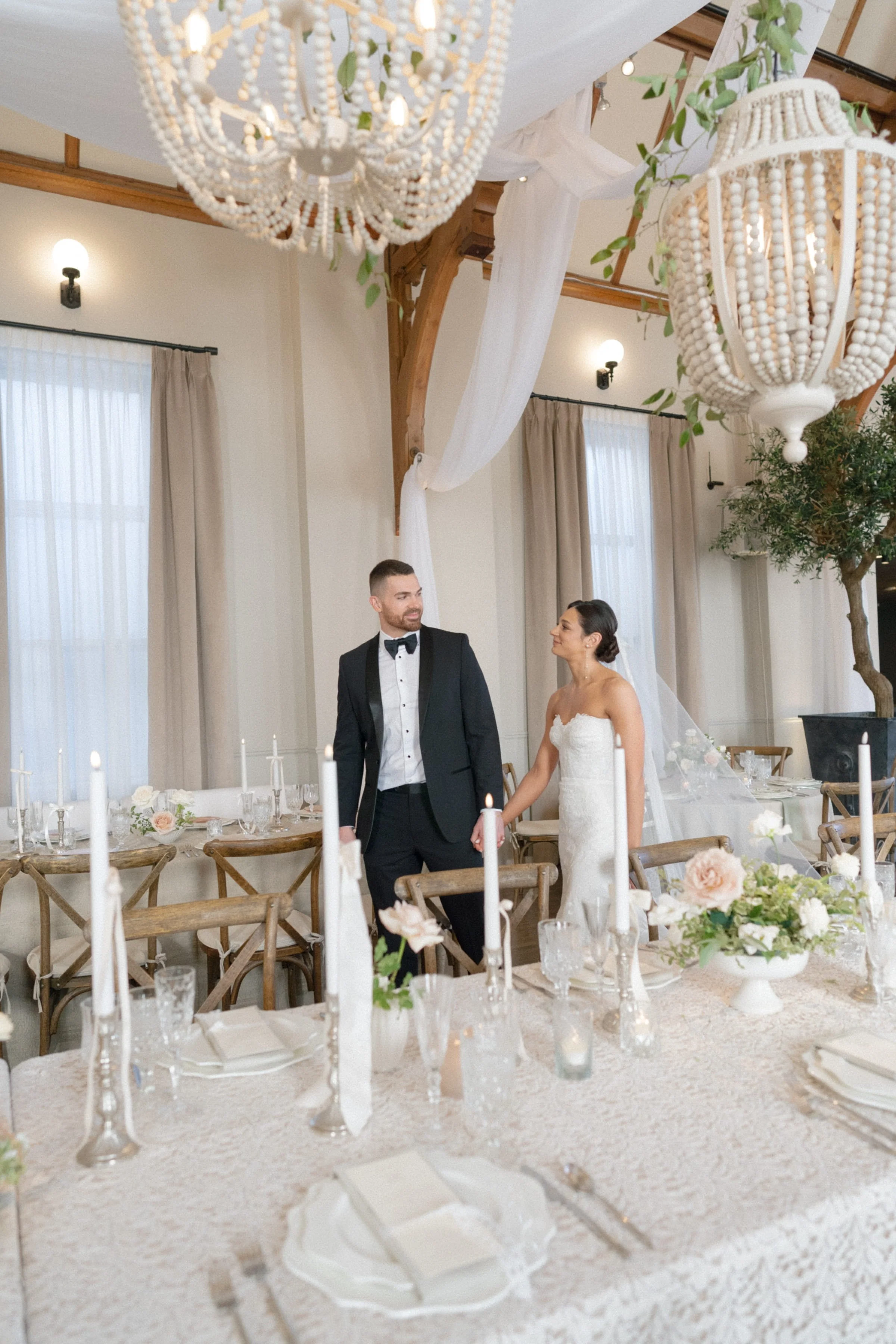 A bride and groom holding hands during their wedding reception, decorated with hanging lanterns, floral arrangements, candles, and tables with white linens.