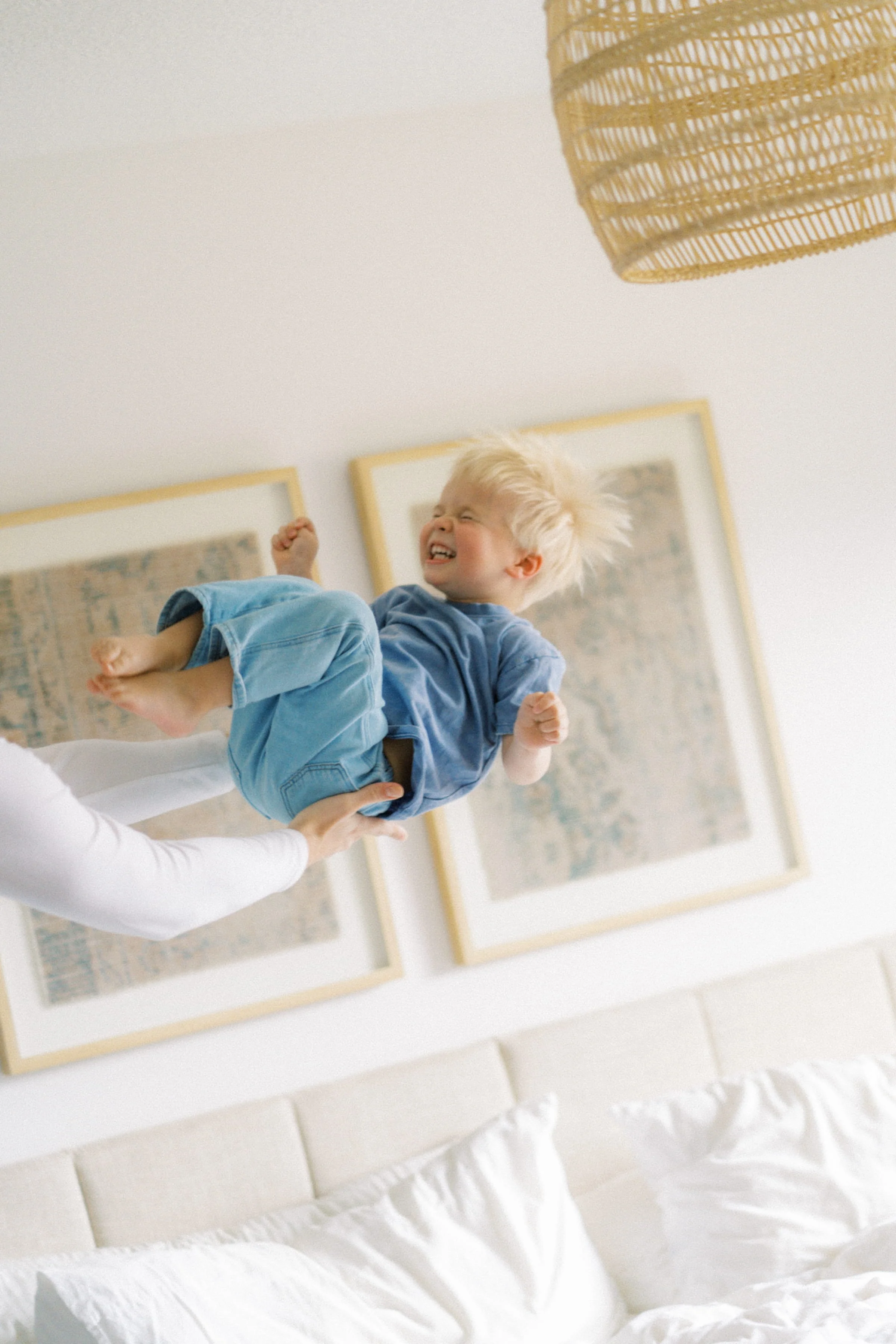 A child with blonde hair, wearing a blue shirt and blue pants, playing and smiling while being lifted in the air by an adult's hand in a bedroom with framed artwork on the white wall.