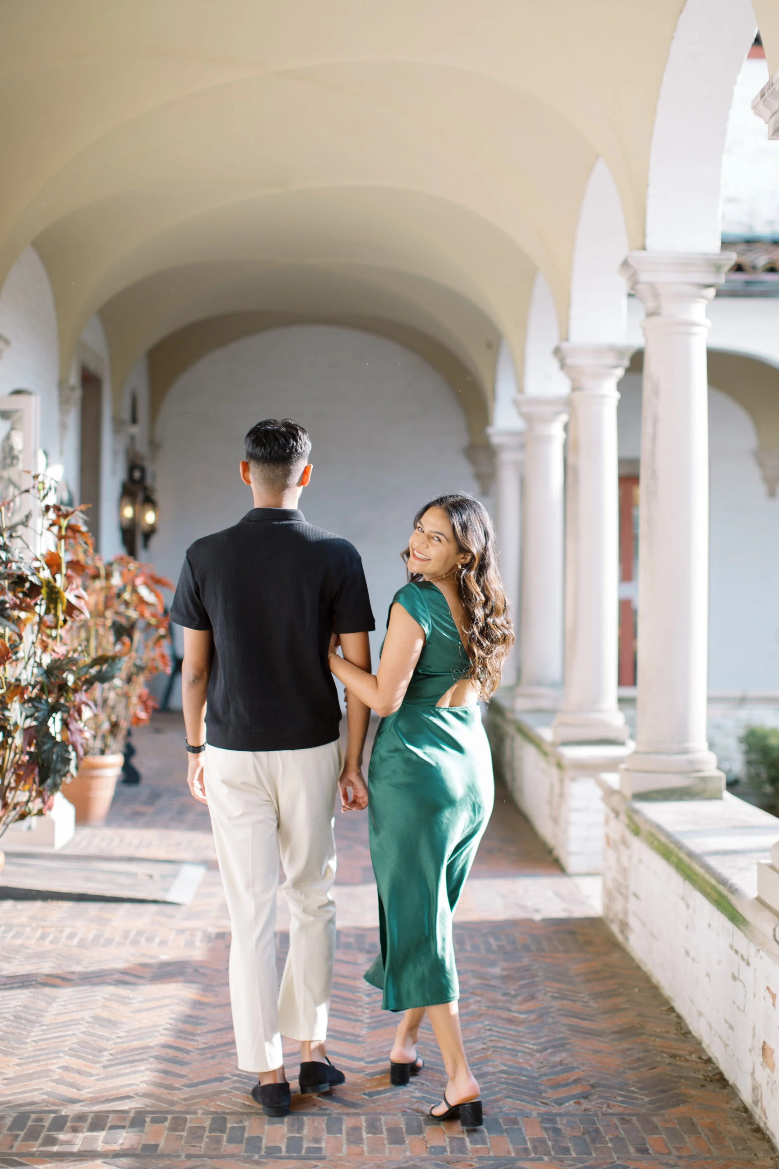 A young woman and a man walking hand in hand under an arched walkway with classic columns. The woman is looking back and smiling, wearing a green satin dress and high heels, while the man is seen from behind, dressed in a black shirt and white pants.
