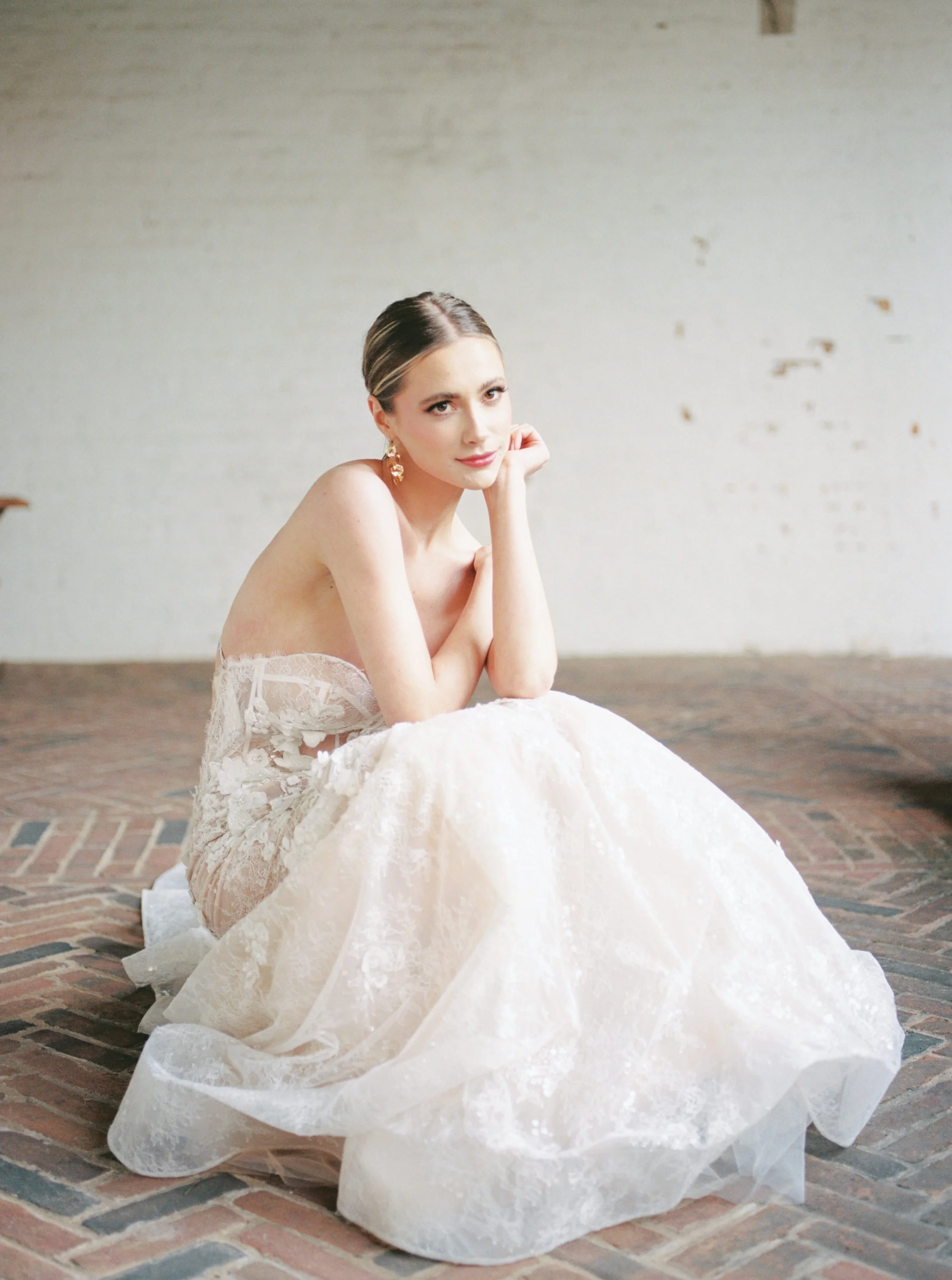 A woman with dark hair in an elegant updo wearing gold earrings and a strapless lace dress, sitting on a brick floor with her chin resting on her hand against a plain wall.