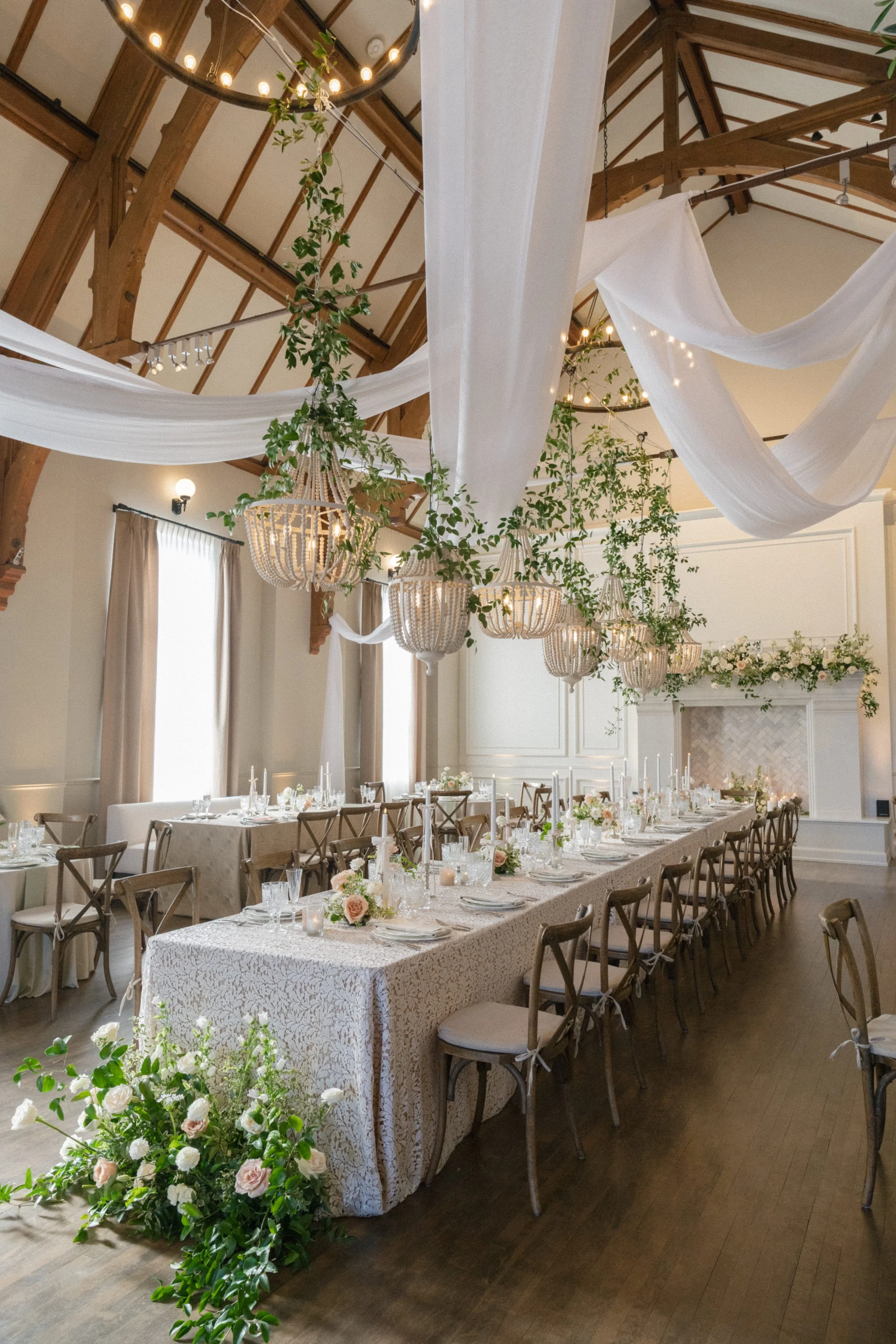 Elegant indoor wedding reception setup with a long table decorated with pink and white floral arrangements, tableware, and candles, adorned with white lace tablecloth and surrounded by wooden chairs. Drape and greenery decorations hang from the ceiling, which features wooden beams and chandeliers, in a bright room with large windows and beige curtains.