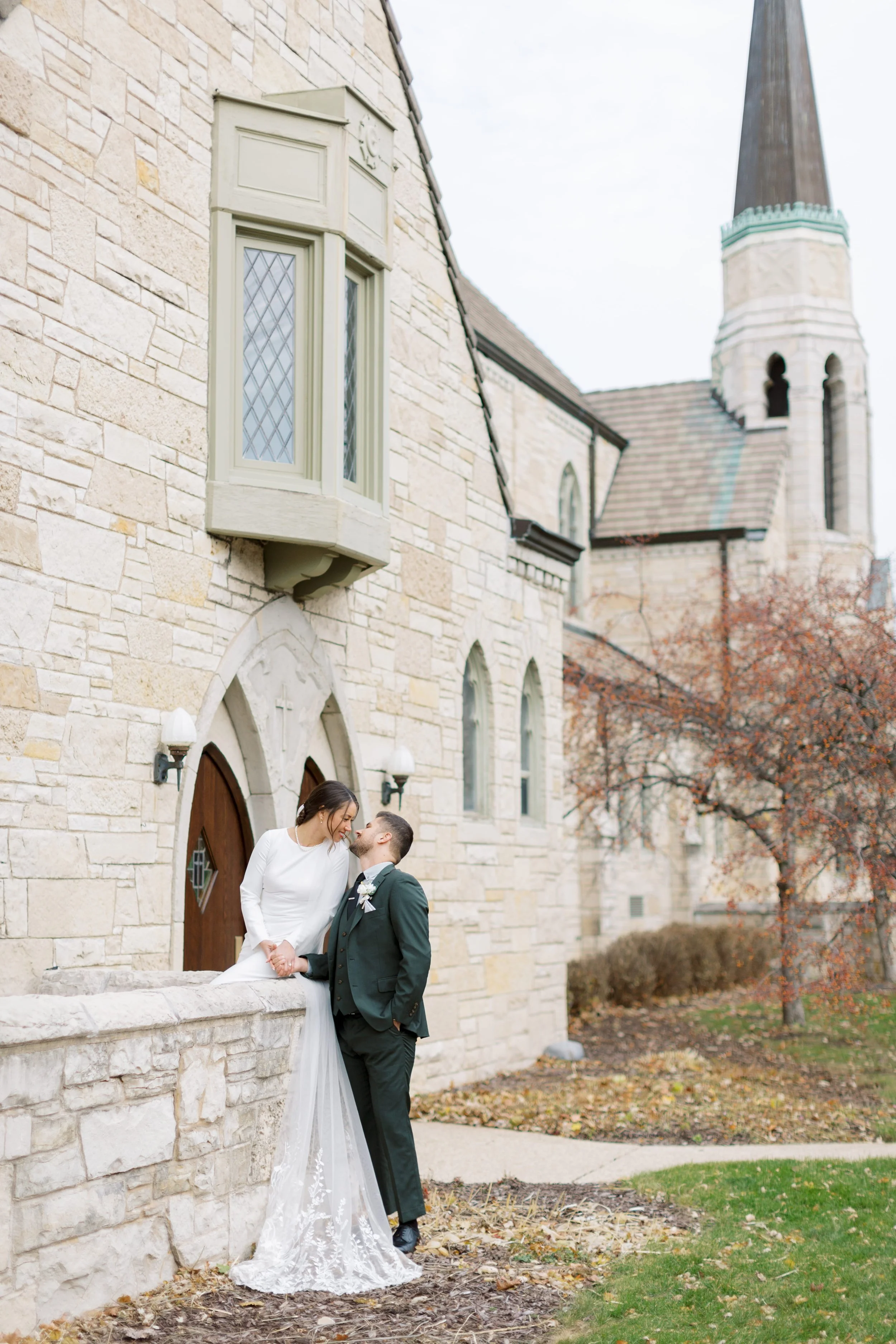 A bride and groom pose outside a stone church, holding hands and smiling at each other on a fall day with trees shedding leaves.