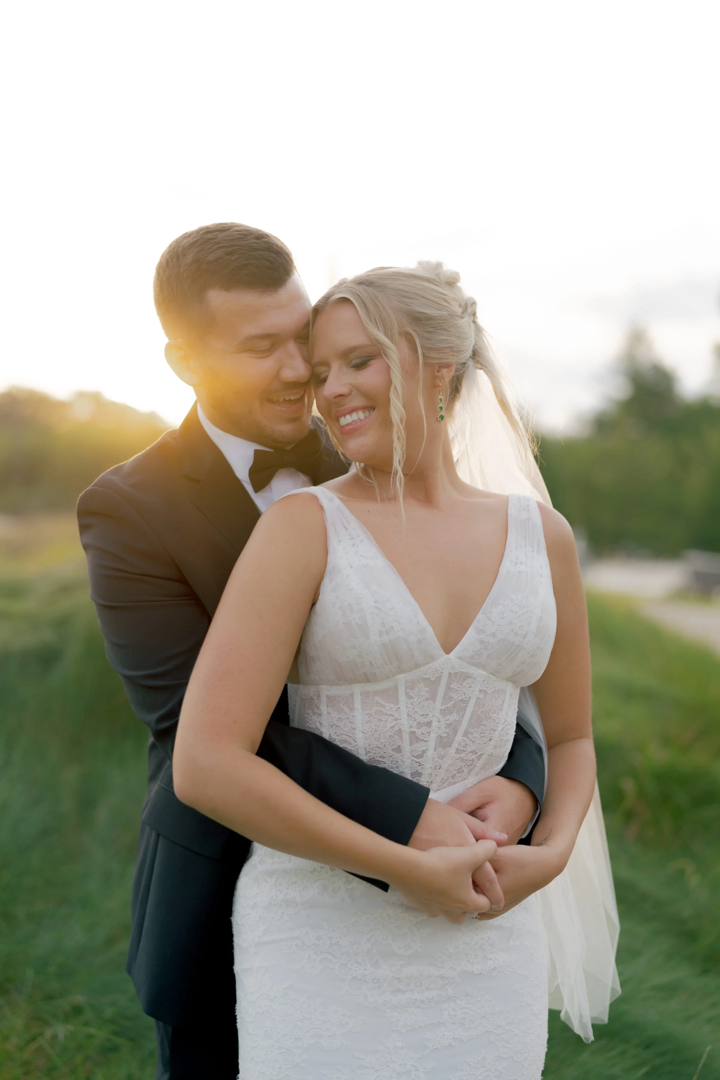 A newlywed couple smiling and embracing outdoors in a grassy field at sunset.