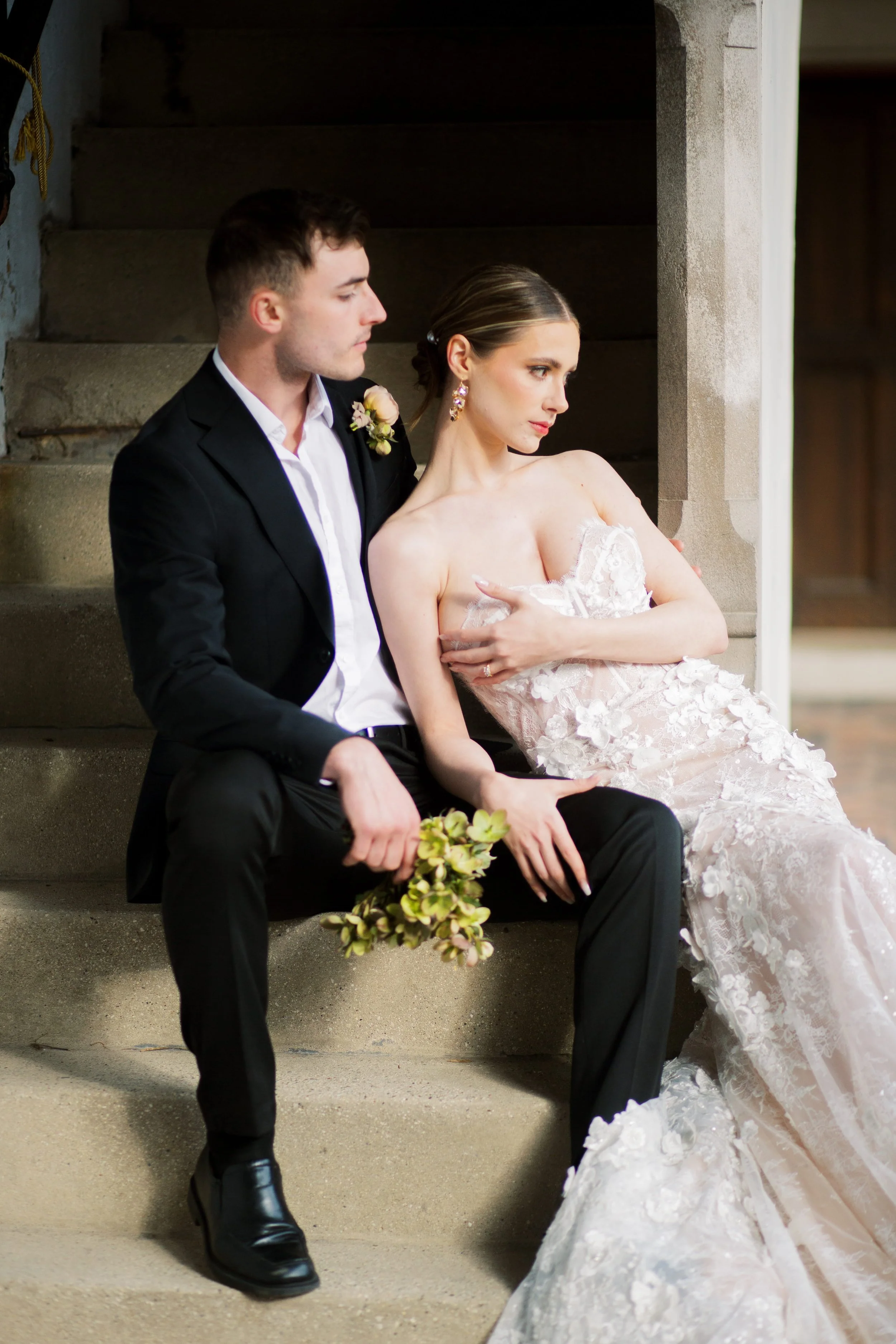 A man in a black suit and a woman in a white lace wedding gown sitting on concrete stairs, the man holding a bouquet of flowers and the woman looking away.