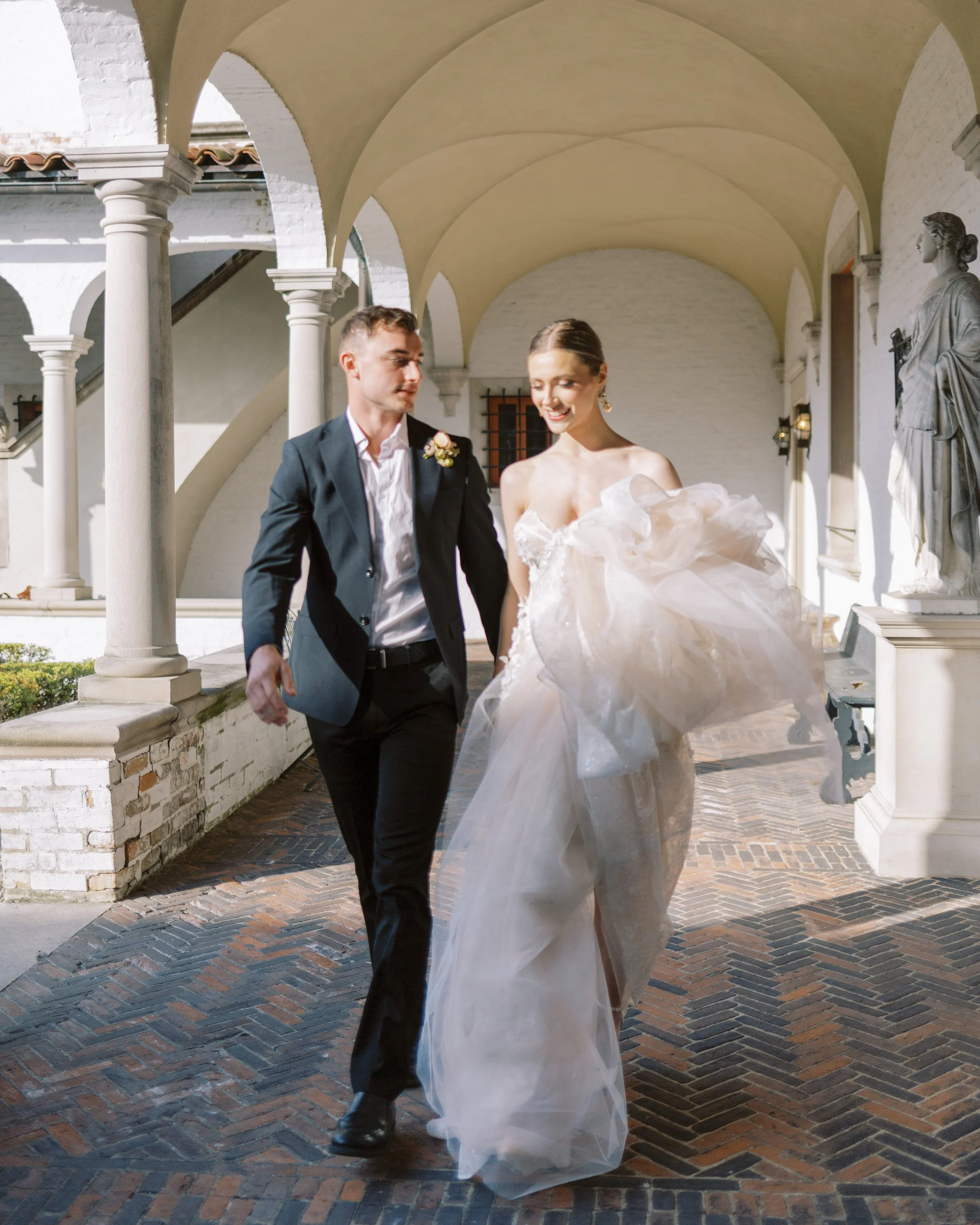 A bride and groom walking together outside under a covered walkway with arched ceilings, white brick walls, and decorative columns, smiling and looking happy after their wedding.