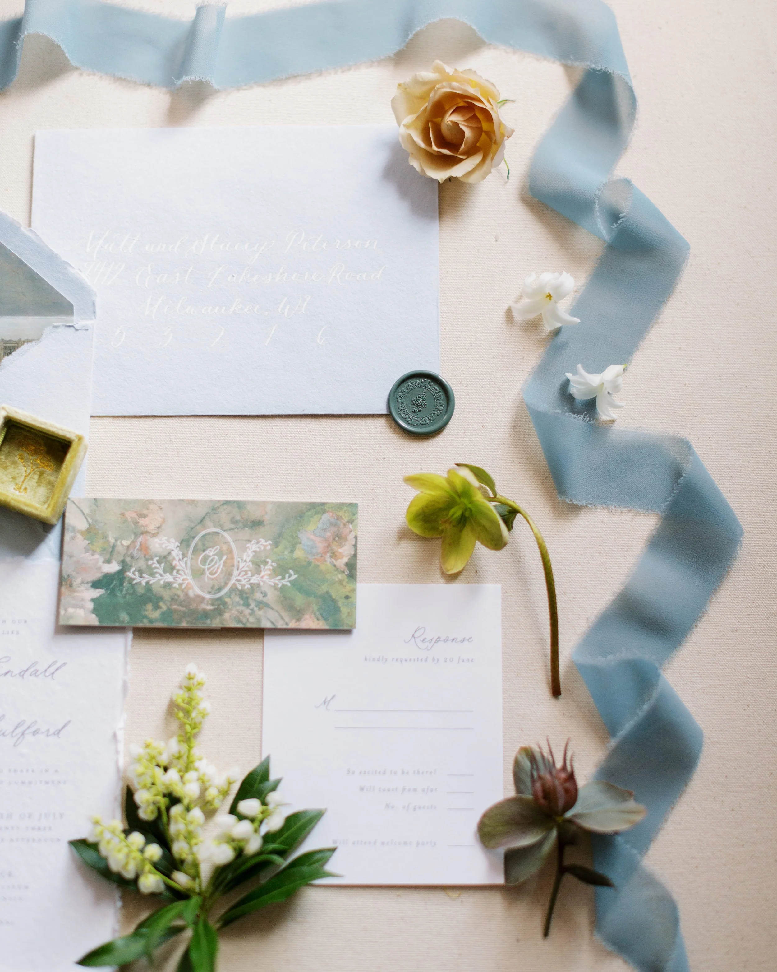 A wedding invitation suite is displayed on a light fabric background, embellished with flowers, a blue ribbon, and a wax seal. The invitation includes elegant calligraphy and botanical illustrations.