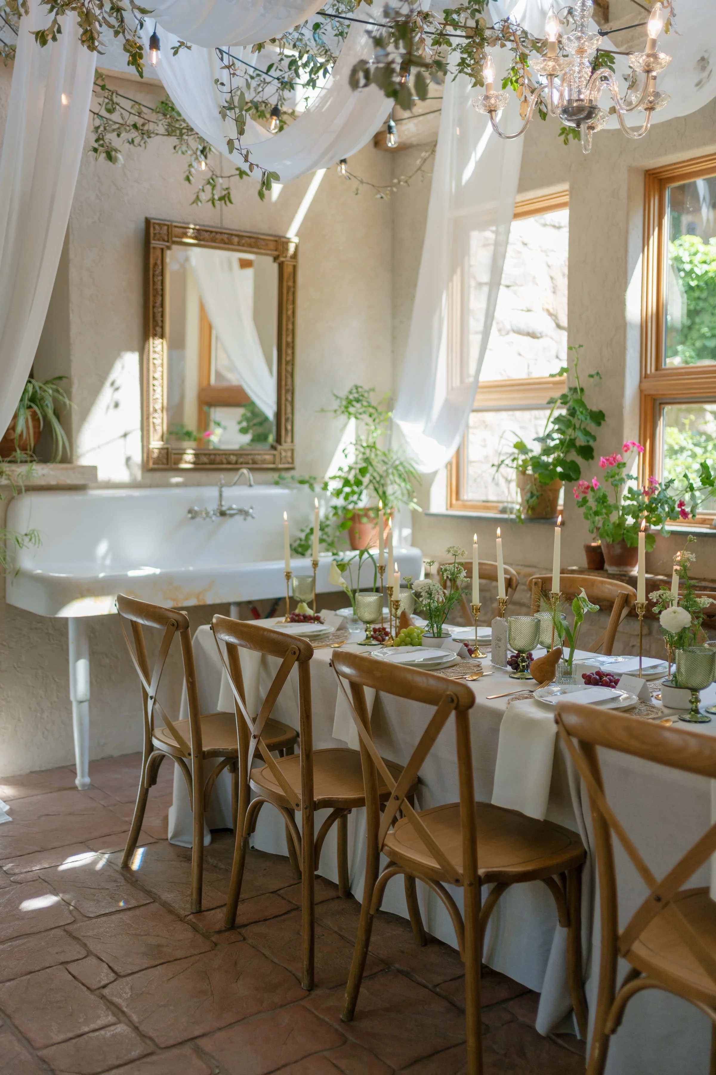 A dining room decorated for a celebration with a long table covered in a white tablecloth, set with plates, glasses, candles, and floral centerpieces. Wooden chairs surround the table. The room has large windows, stone walls, potted plants, and draped white fabric with hanging greenery and a chandelier.