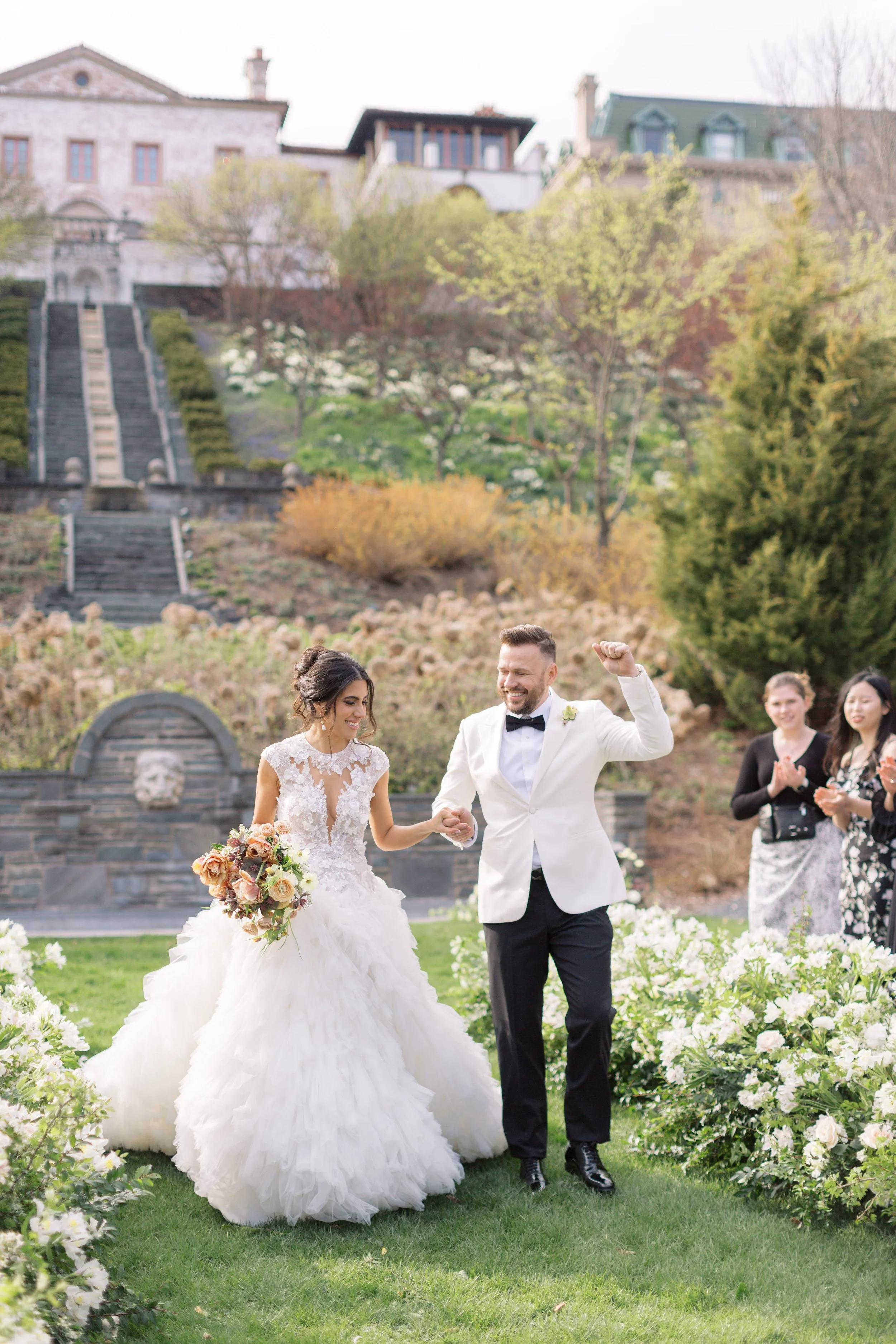 A bride and groom celebrating their wedding outdoors on a grassy lawn, surrounded by floral decorations and white flowers, with family and friends in the background and a landscaped hillside with trees and houses behind them.
