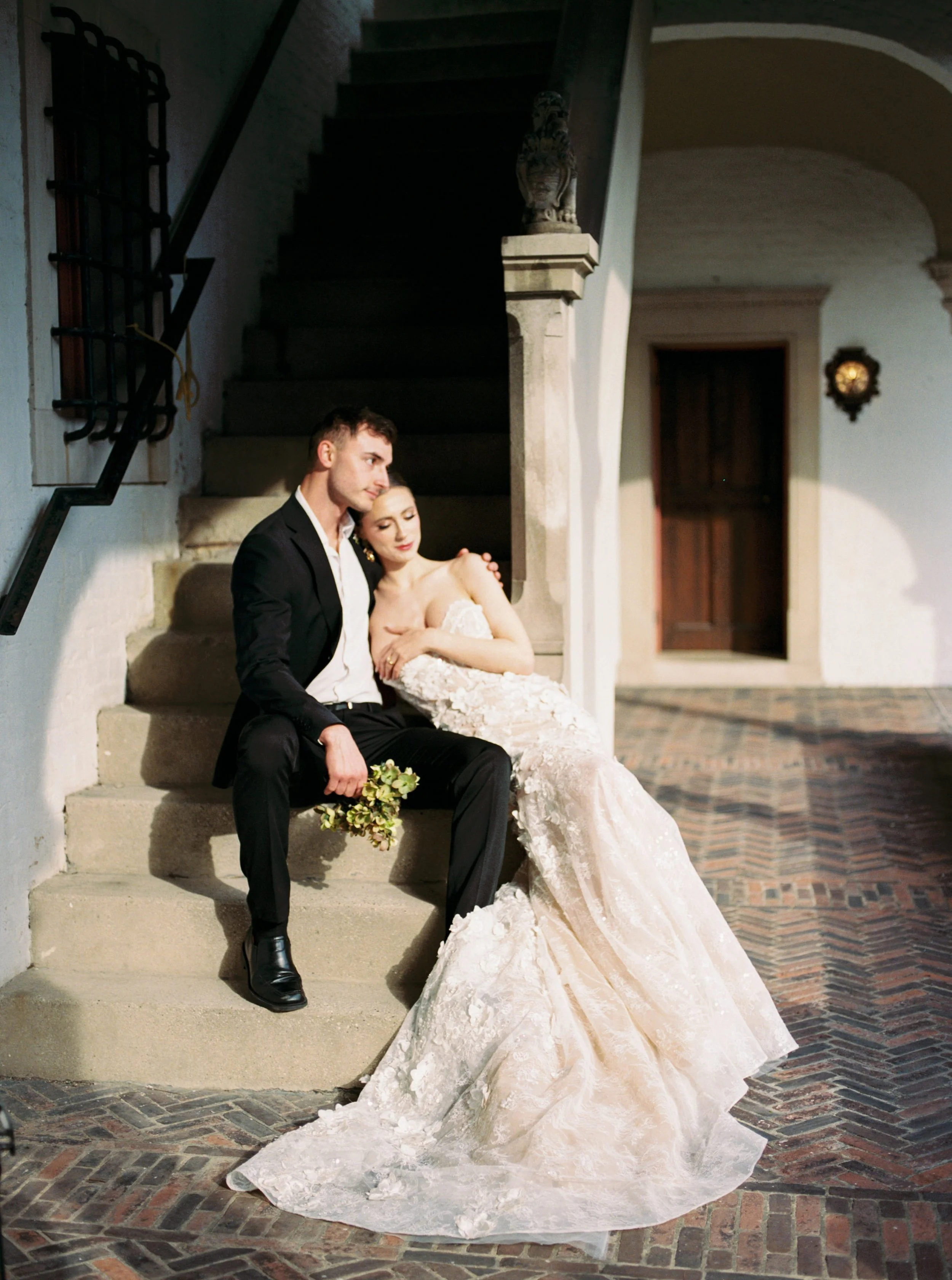 A bride and groom sit on stairs inside a rustic building. The groom wears a black suit and the bride in a white wedding gown holds a small bouquet. The bride rests her head on the groom's shoulder with her eyes closed. The setting includes stone steps, a decorative crest on a column, and warm interior lighting.