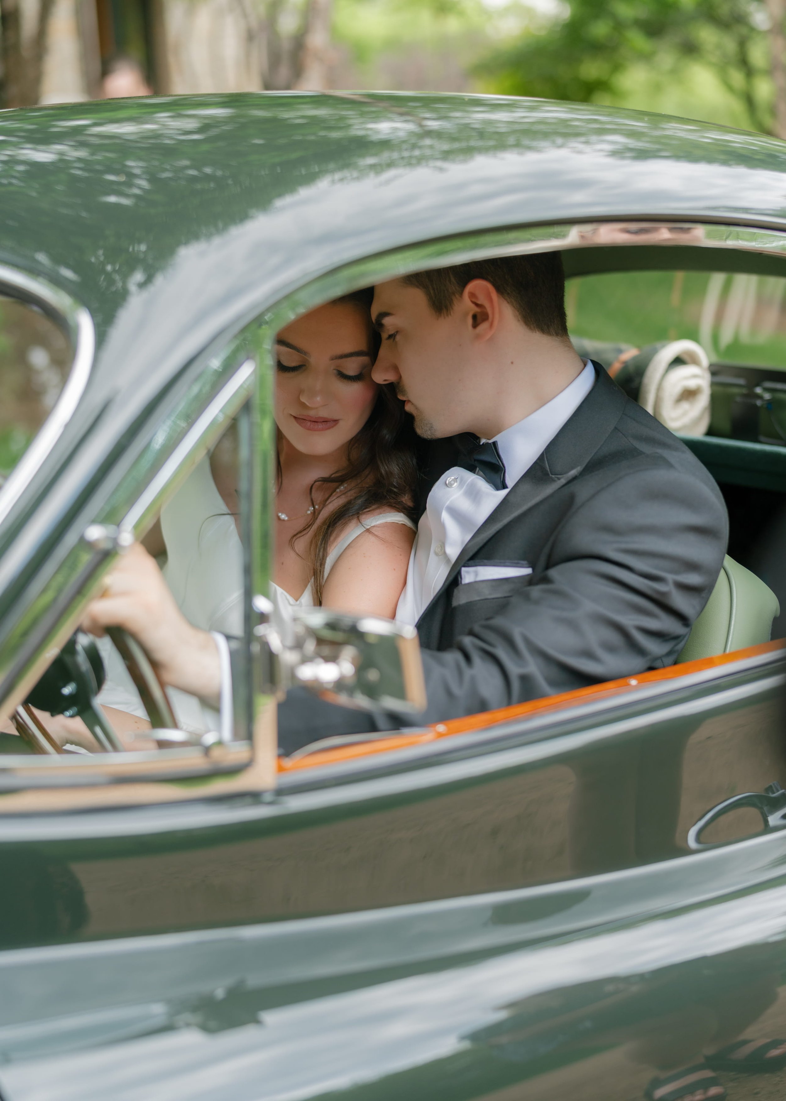 A couple dressed in formal wedding attire sitting close inside a vintage green car, leaning their foreheads together with eyes closed, surrounded by a nature background.