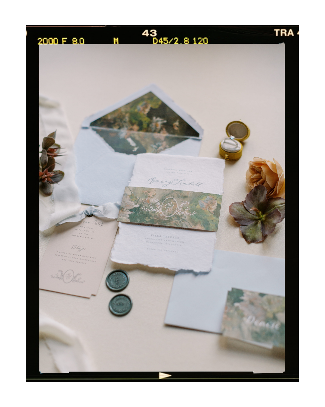 Flat lay of wedding stationery and accessories, including invitation cards, envelopes, wax seals, a floral pocket, and a jewelry box, arranged on a light-colored surface with decorative flowers.