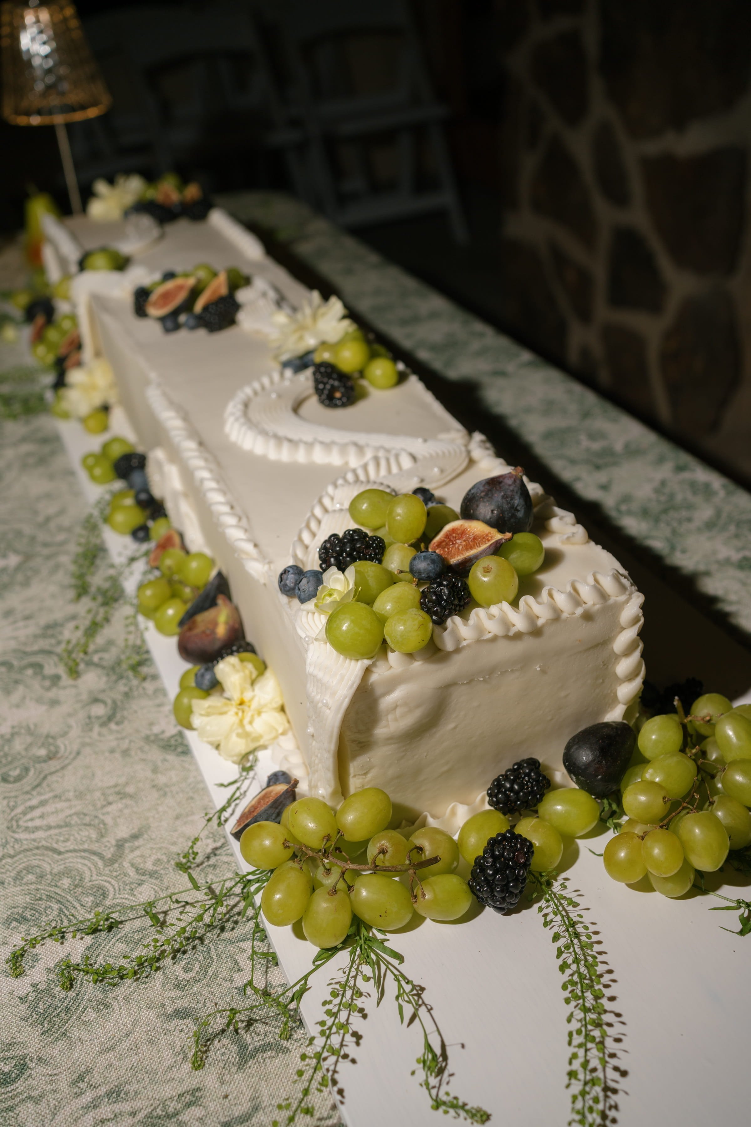 A long rectangular white cake decorated with green grapes, blackberries, figs, and white flowers, placed on a patterned tablecloth with greenery around the base.
