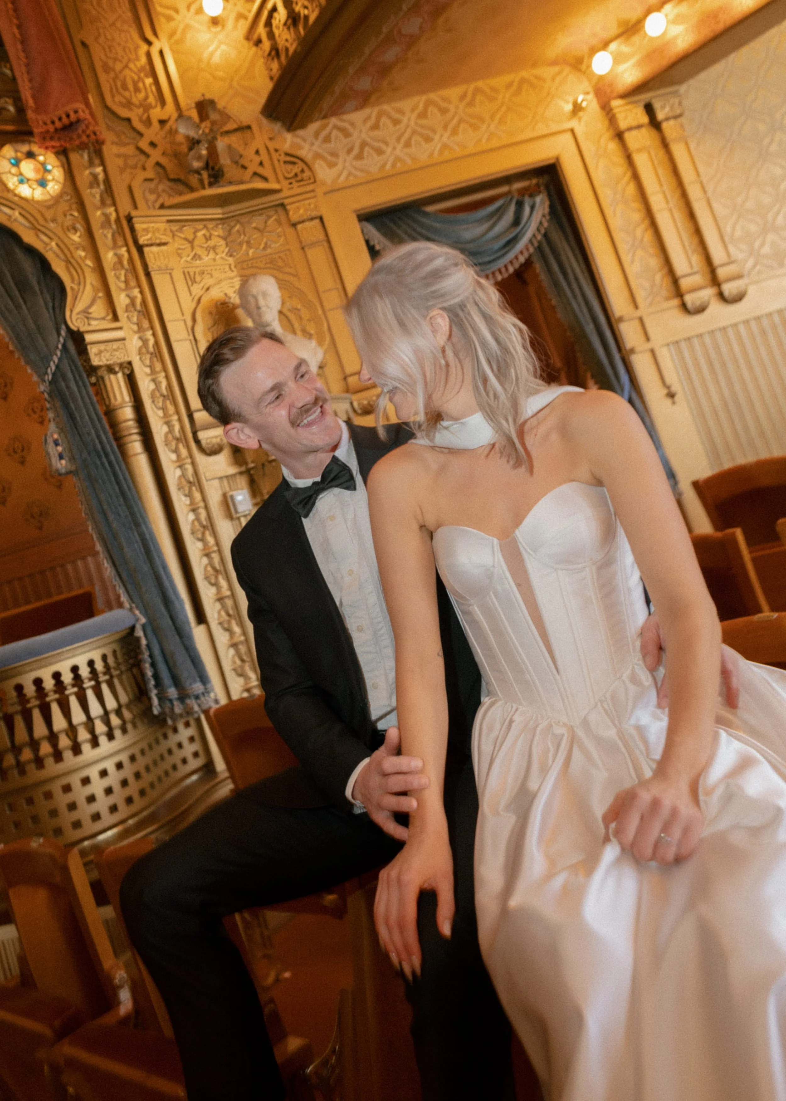 A smiling man in a tuxedo and bow tie holding hands with a woman in a white strapless dress, in an ornate gold-colored room with classical decor and a bust sculpture.