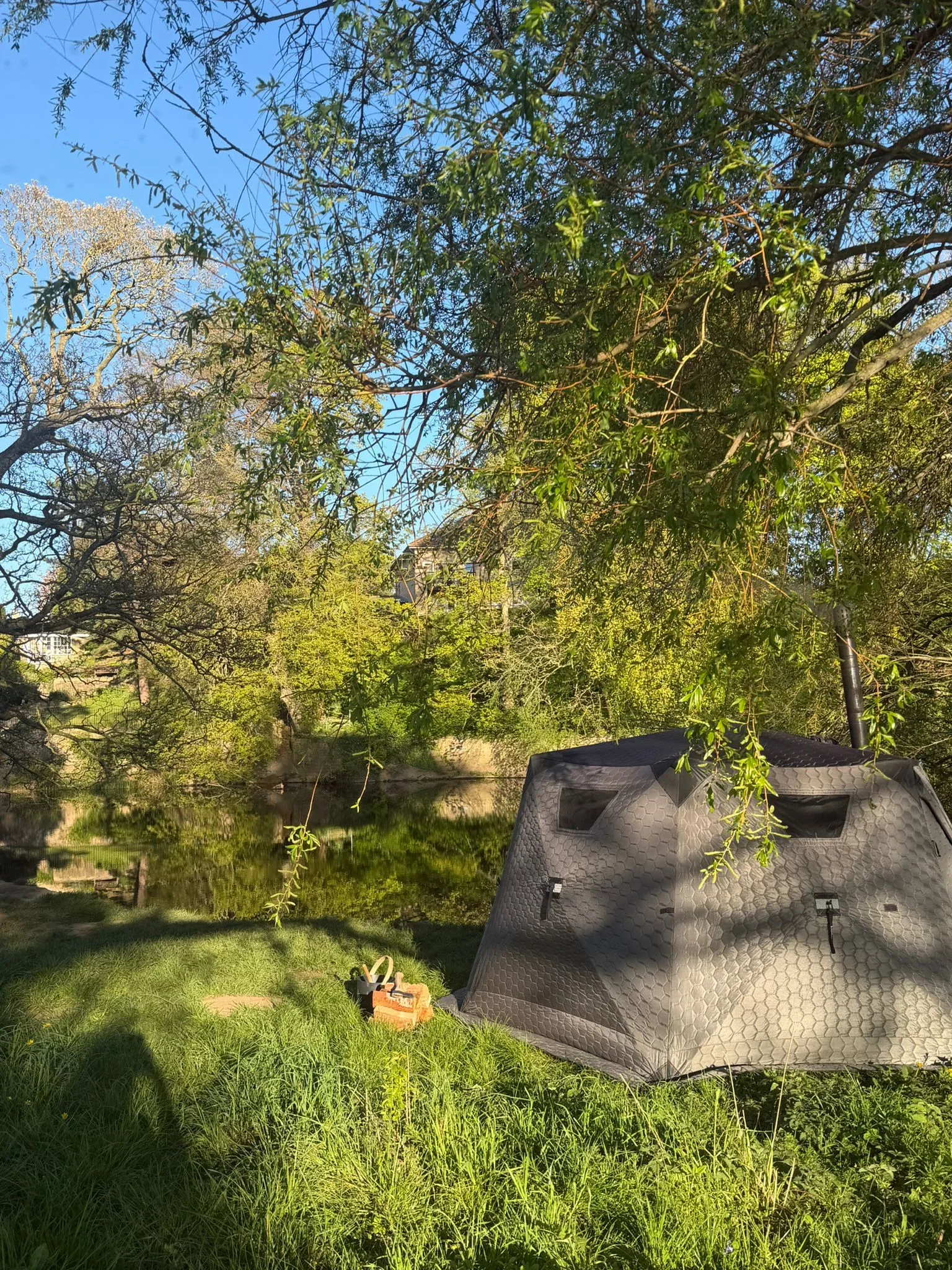 A gray pop-up camper tent set up on a green grassy area near a river with trees in the background. The sky is blue with some tree branches hanging down in the foreground.