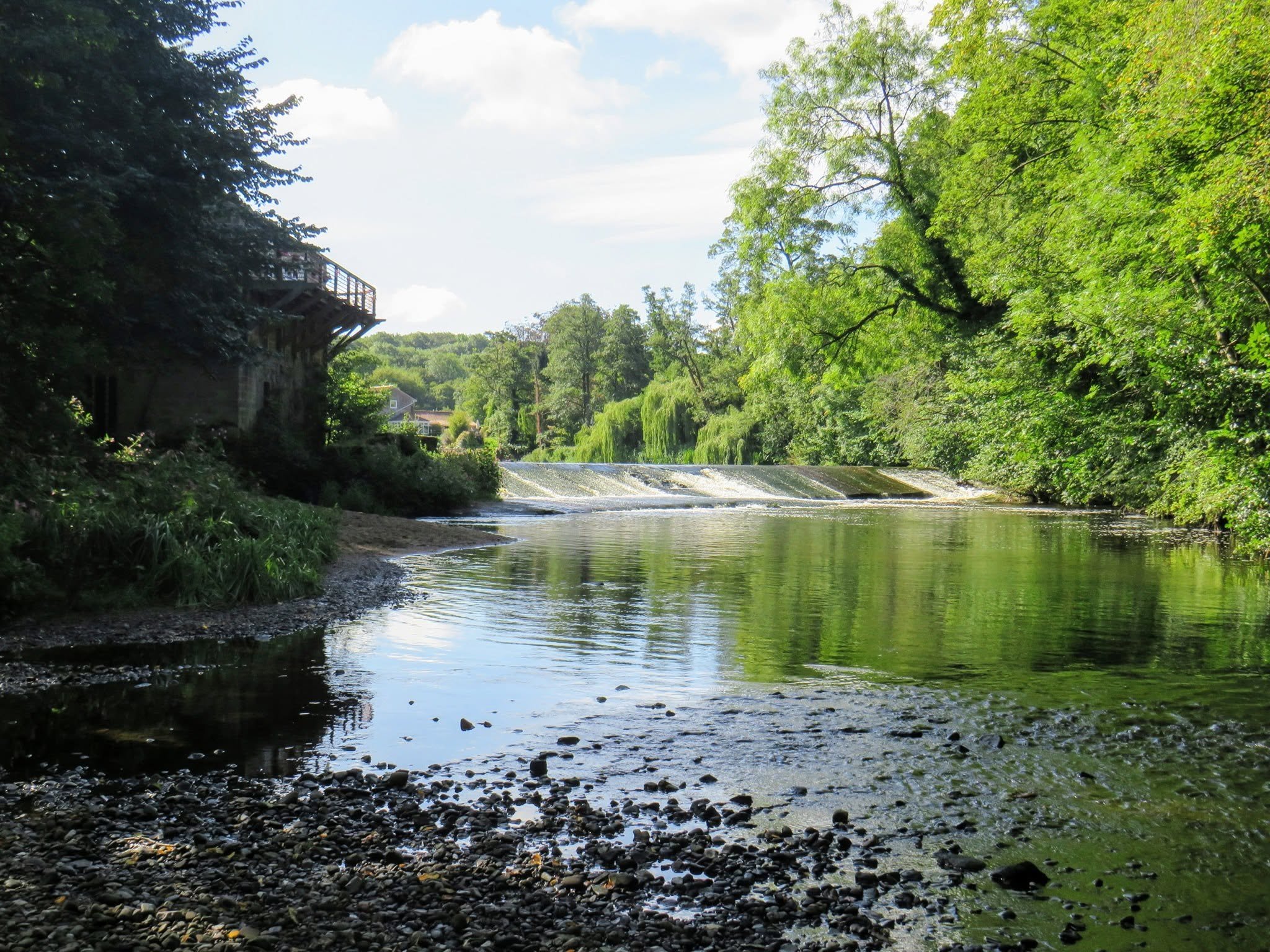 A peaceful river scene with a small waterfall, lush green trees on both sides, and houses visible in the background under a partly cloudy sky.