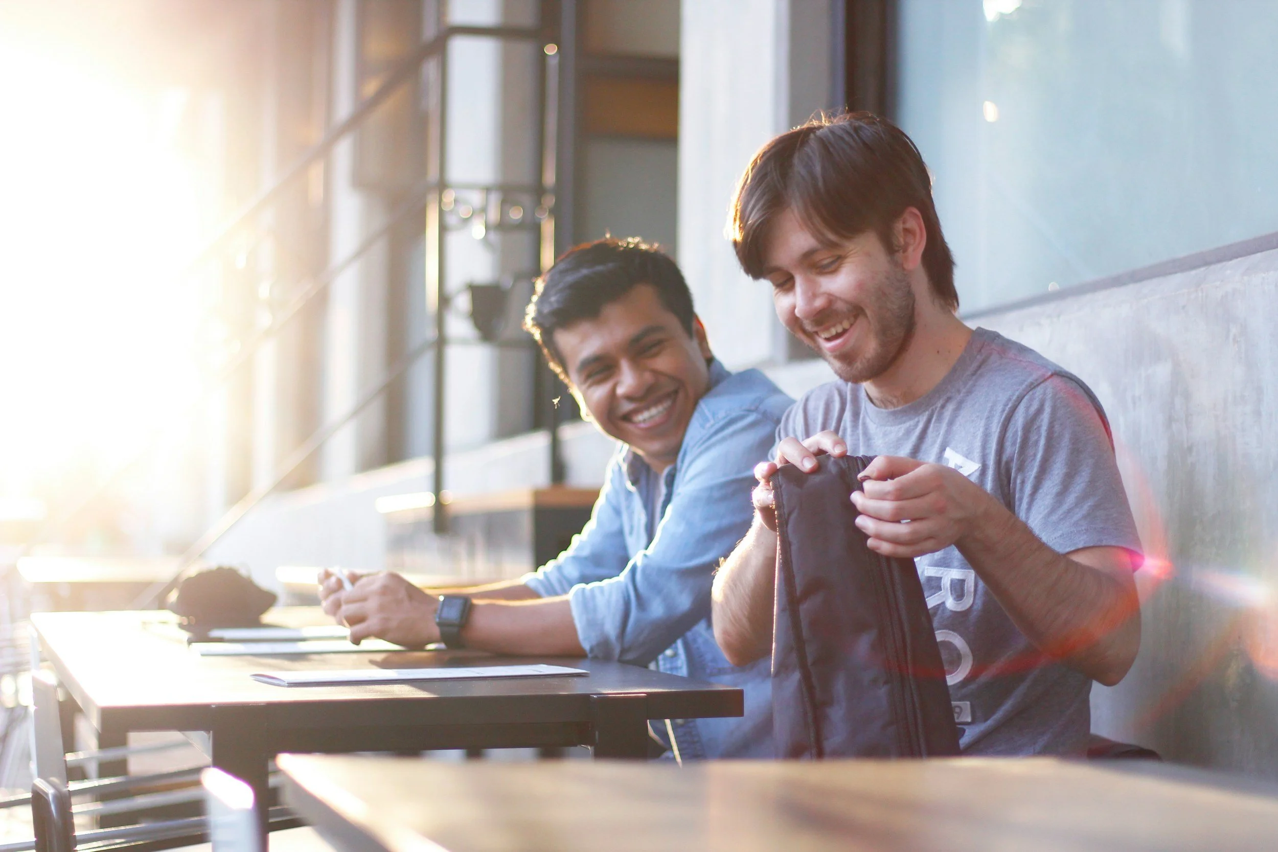Two young men sitting at a table, smiling and in an outdoor space with natural sunlight.