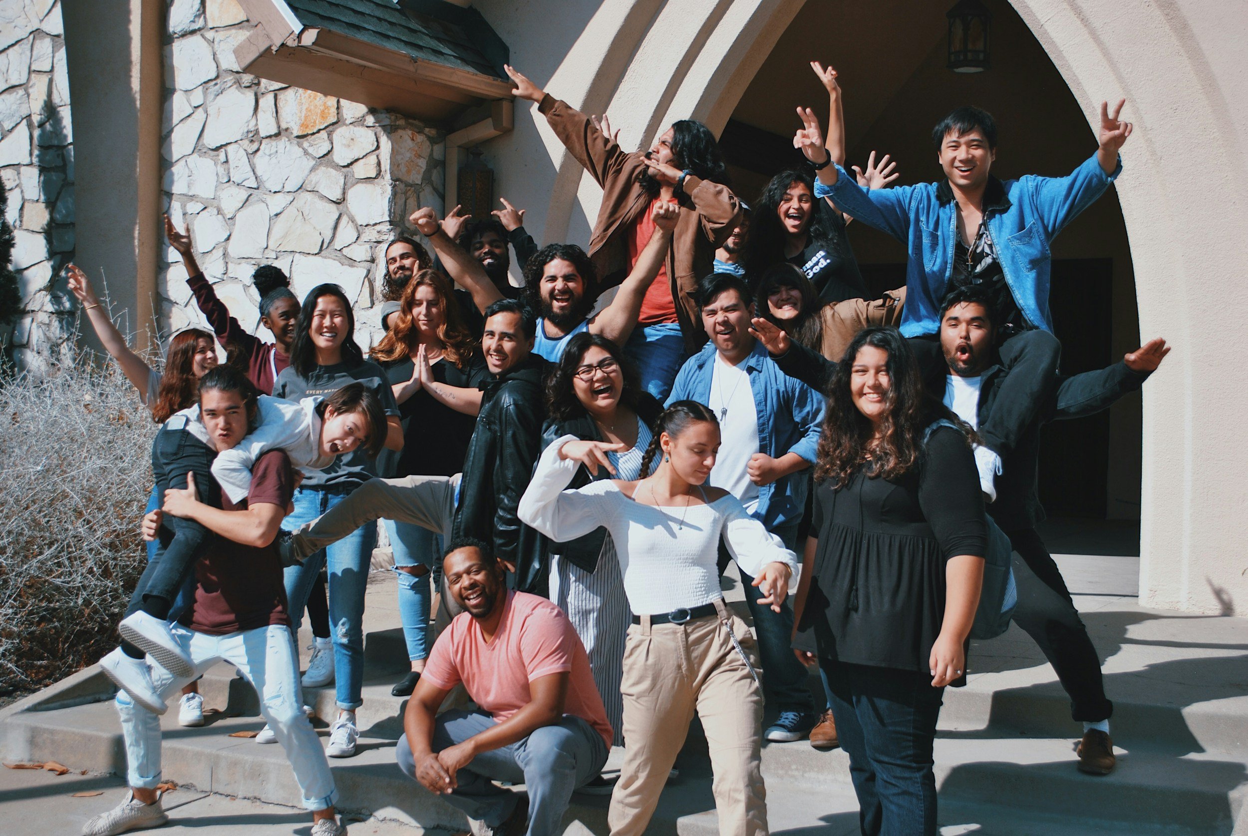 A group of diverse young people standing and sitting on steps outside a stone and stucco church, smiling and making joyful gestures.