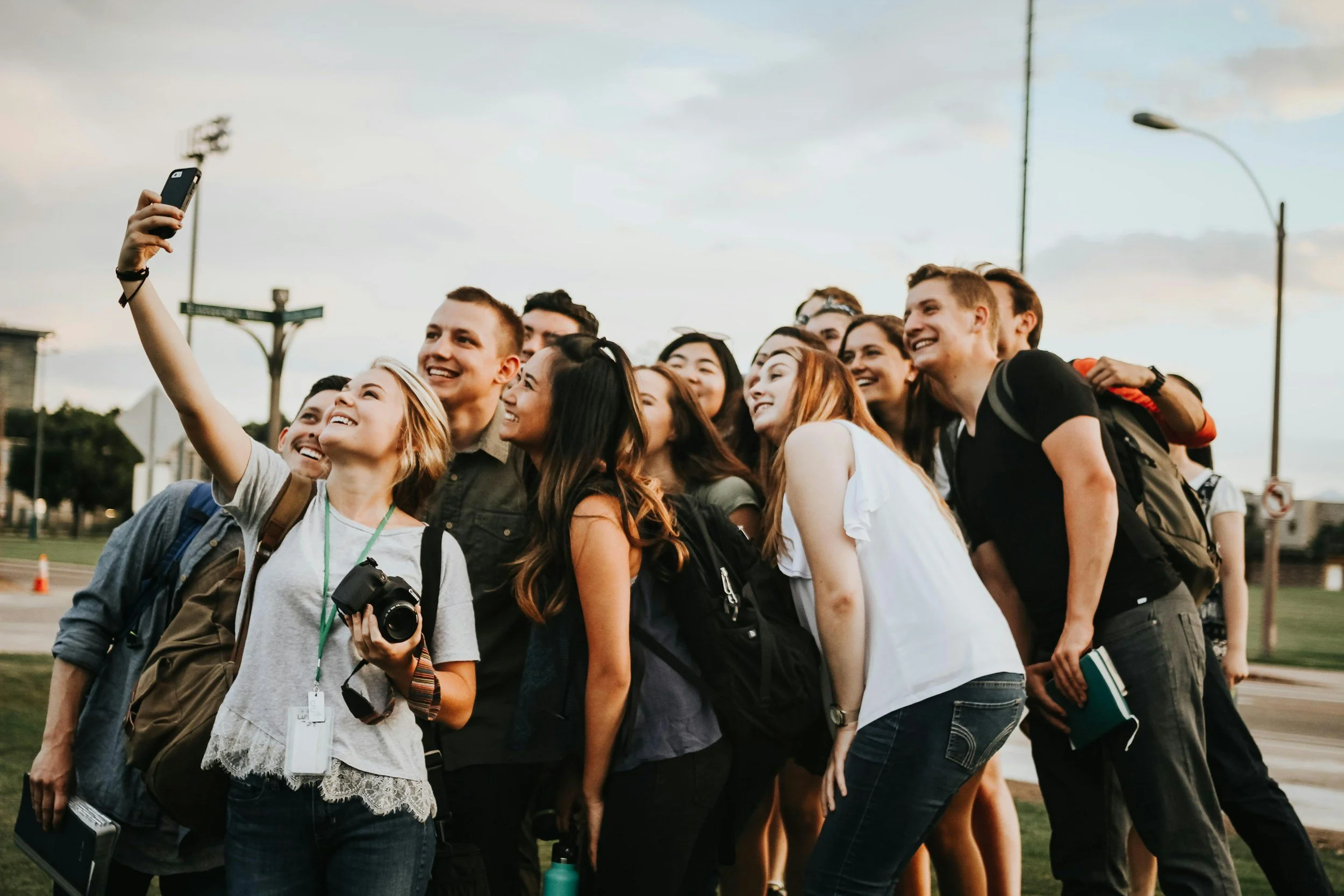 A diverse group of young people taking a selfie outdoors on a field during the daytime, smiling and enjoying themselves.
