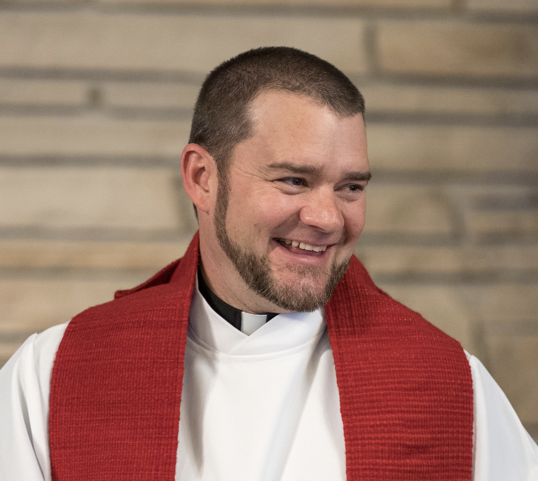 A smiling man wearing a clerical collar and a red stole, standing in front of a wooden background.