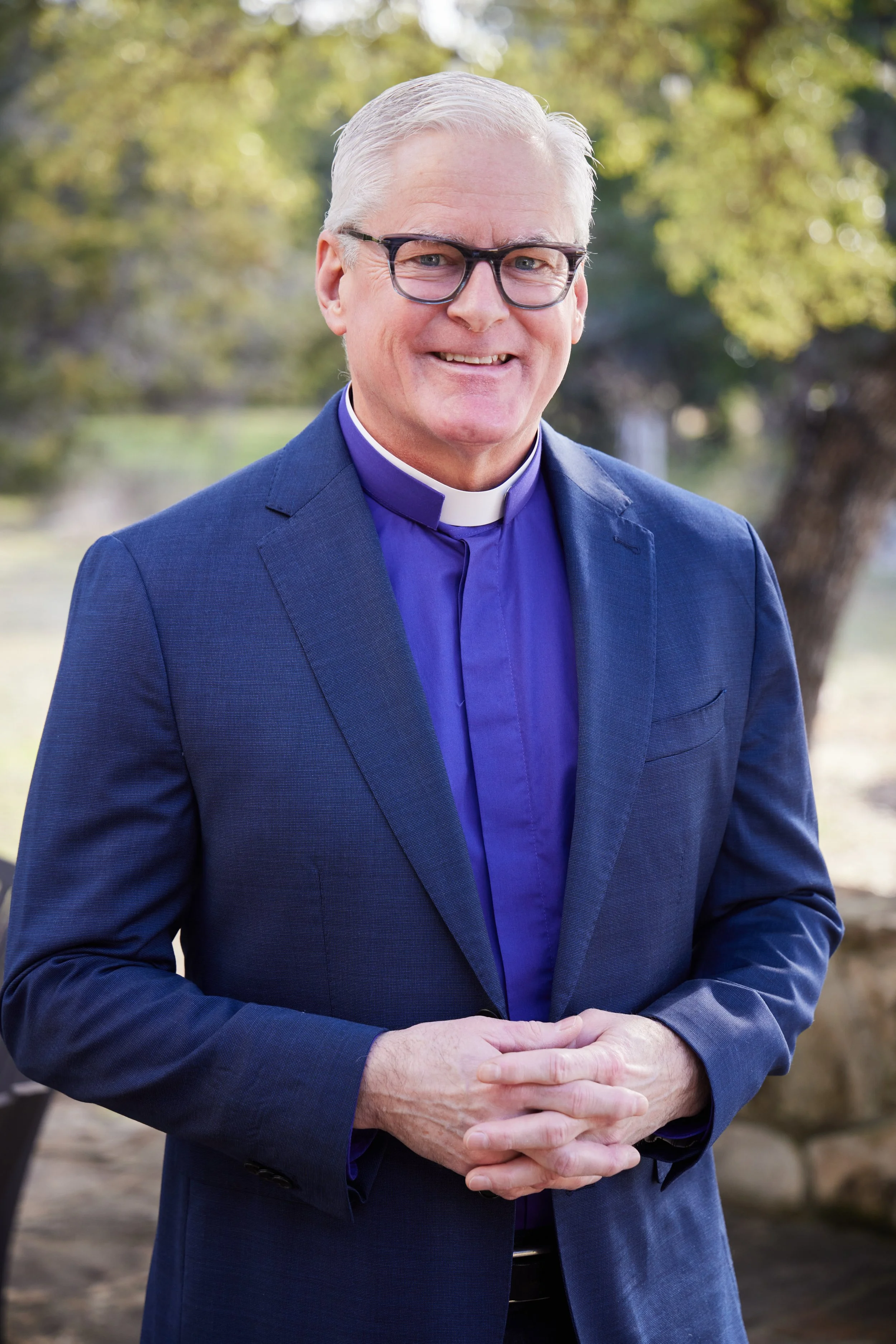 A middle-aged man wearing a navy blue suit, a clerical collar, and glasses standing outdoors with trees and blurred nature in the background, smiling with hands clasped.