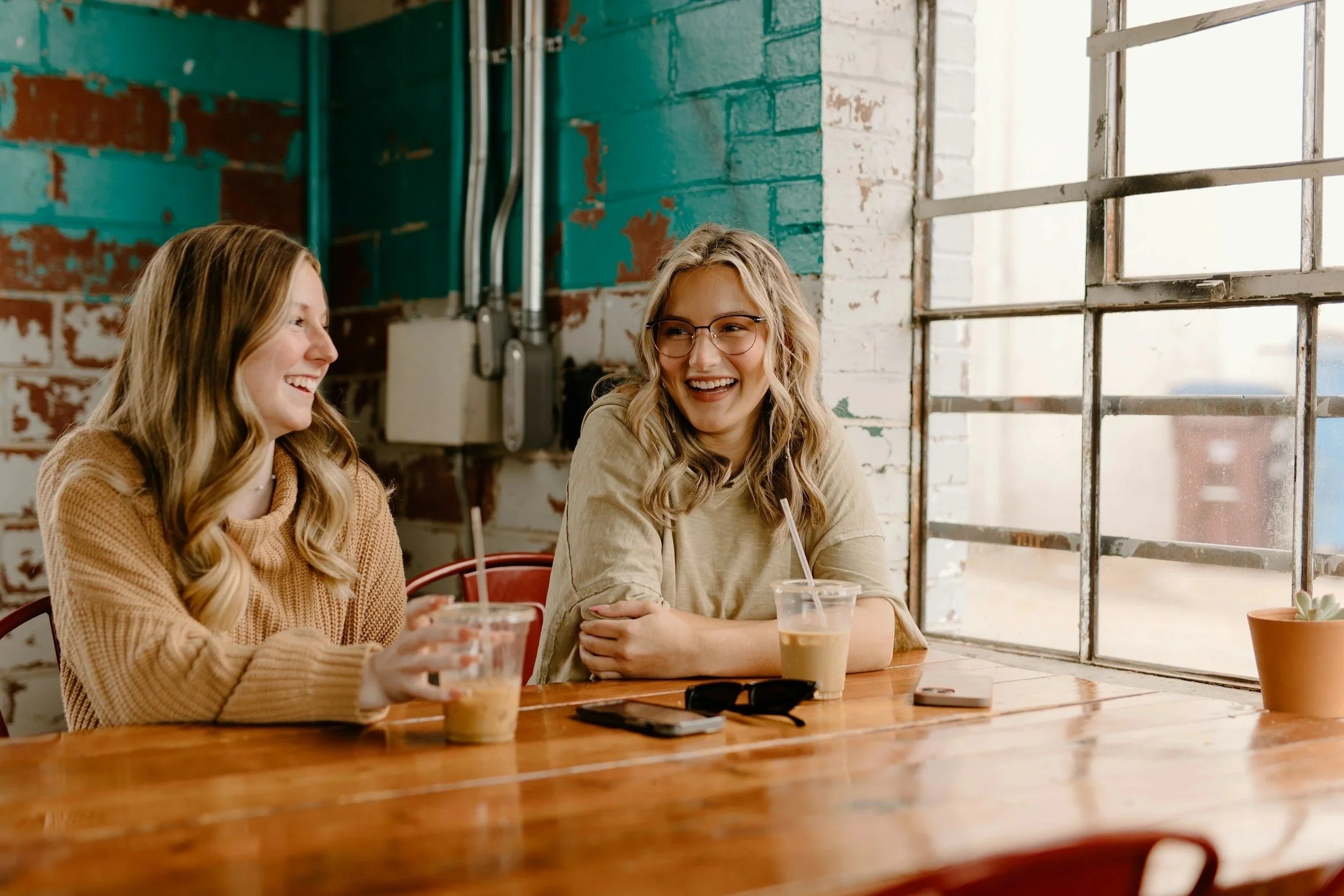Two women sitting at a wooden table in a cafe, smiling and talking, with drinks in front of them, illuminated by large windows with metal frames.