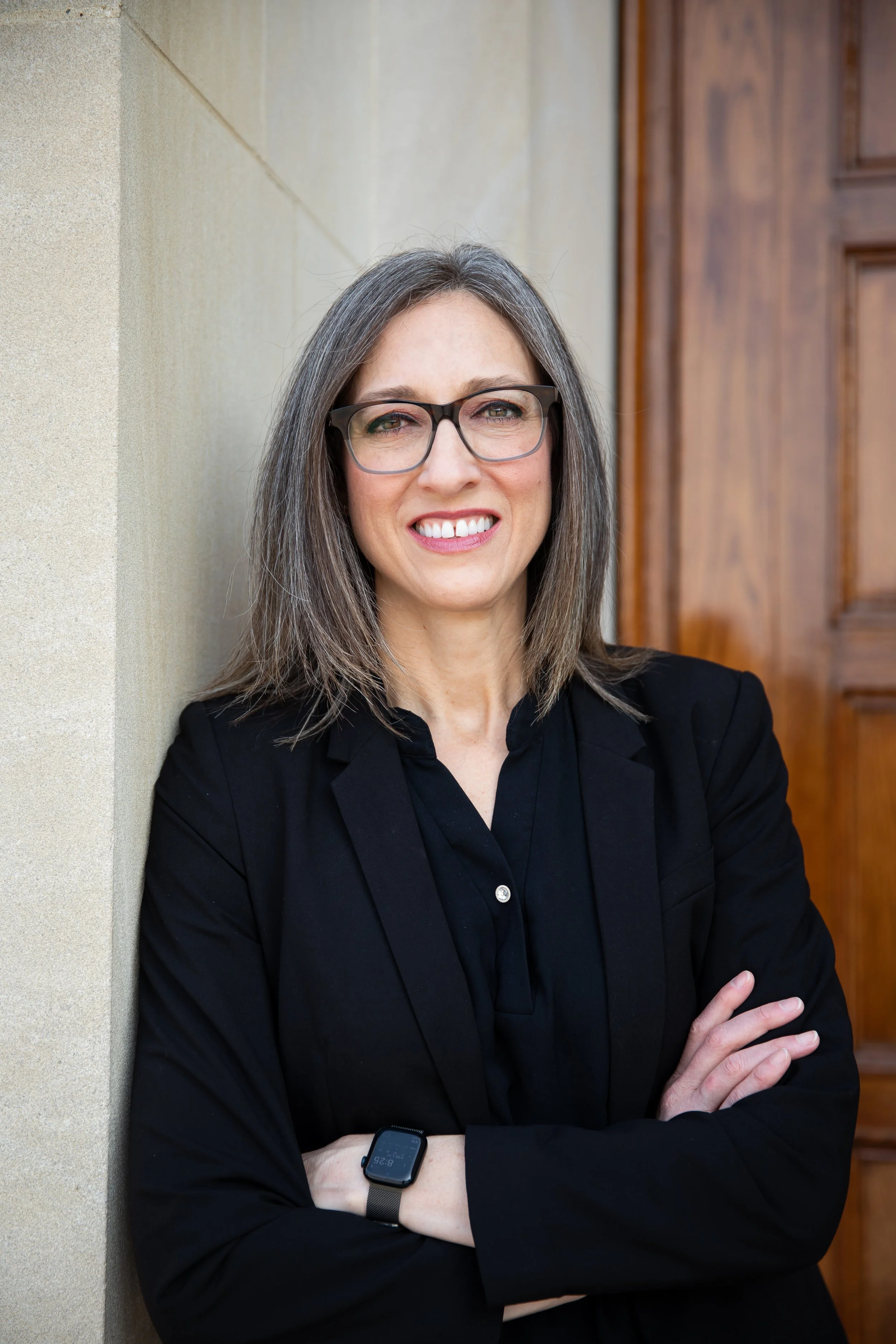 A middle-aged woman with shoulder-length gray hair, wearing glasses, a black blazer, and a smartwatch, standing with arms crossed against a beige wall and a wooden door in the background, smiling at the camera.