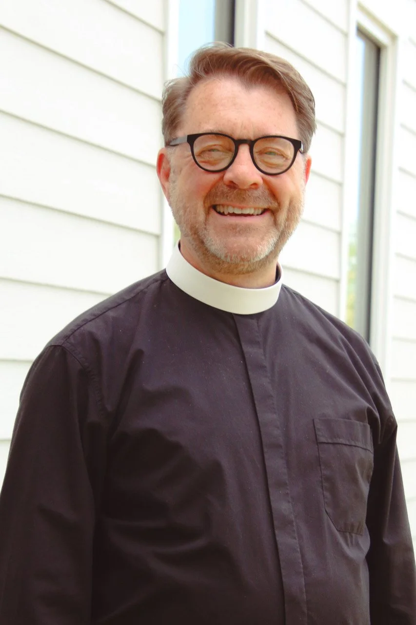 A smiling man dressed as a clergy member, wearing a black shirt with a white clerical collar, standing outdoors near a light-colored building with horizontal siding.