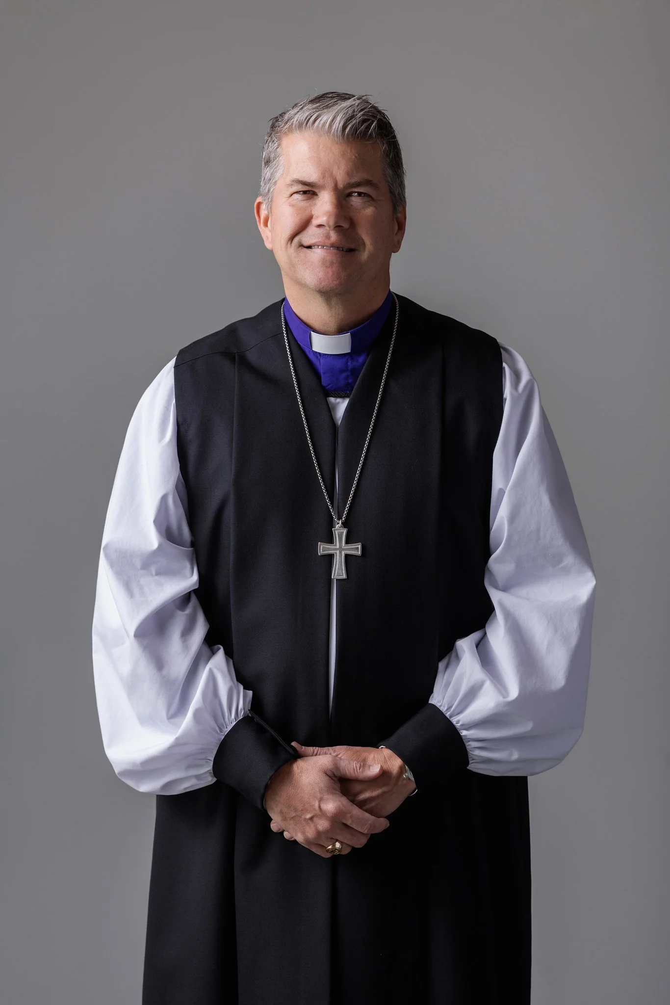 A middle-aged man dressed in clerical attire, wearing a clerical collar and a large cross pendant, standing against a plain gray background.