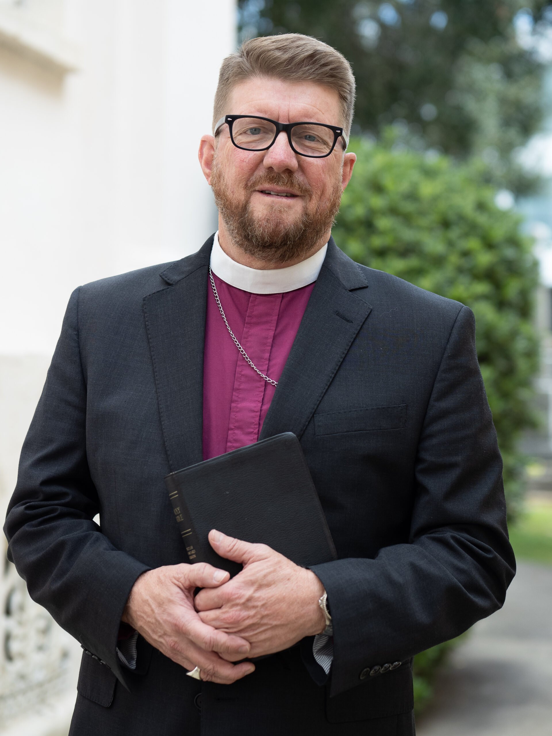 A middle-aged man with glasses, a beard, wearing a black suit and a purple clergy shirt with a white collar, holding a black book, standing outdoors in front of trees.